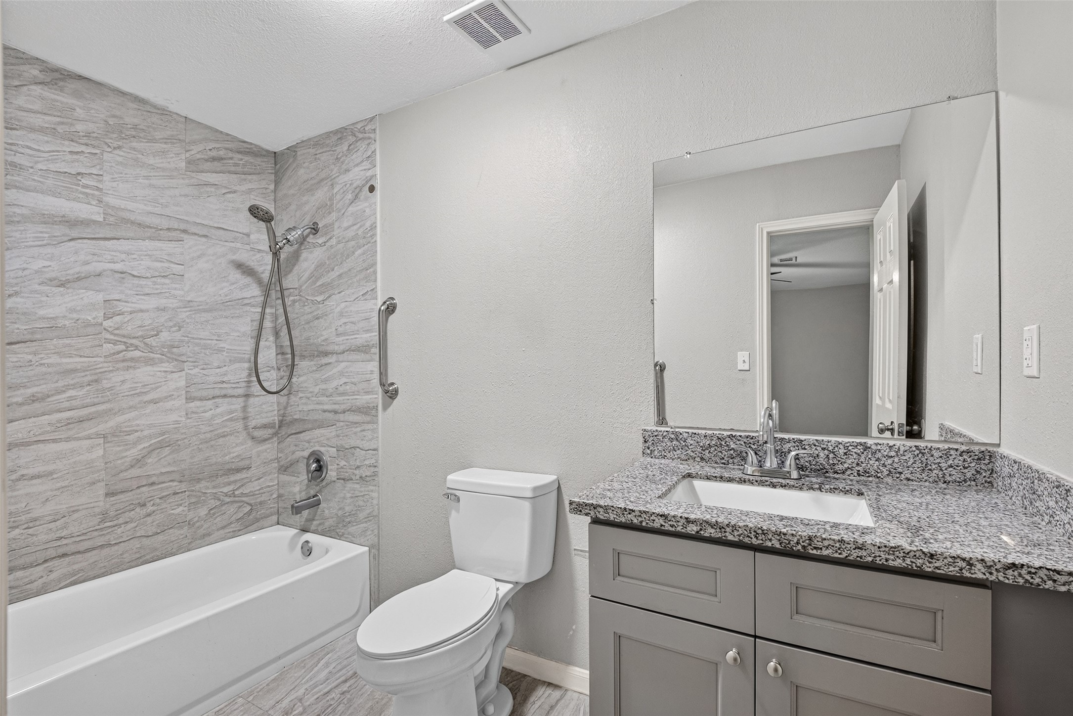 This bathroom features a modern design with gray tones, showcasing a granite countertop vanity, a large mirror, and a sleek bathtub with tiled walls. Perfect for a contemporary home.