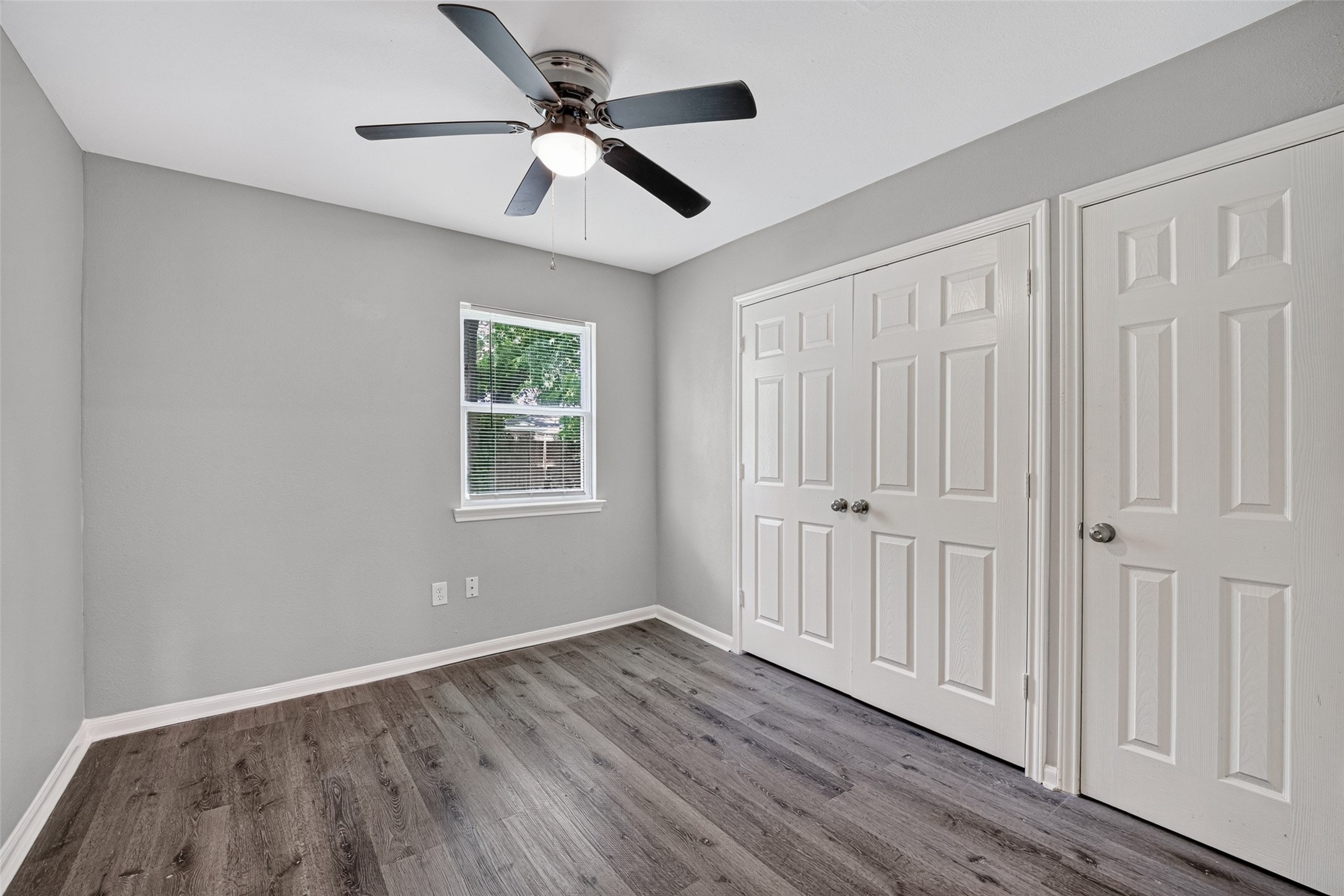 This room features fresh gray walls, dark wood flooring, a ceiling fan, a window for natural light, and double closets with white doors, offering ample storage.