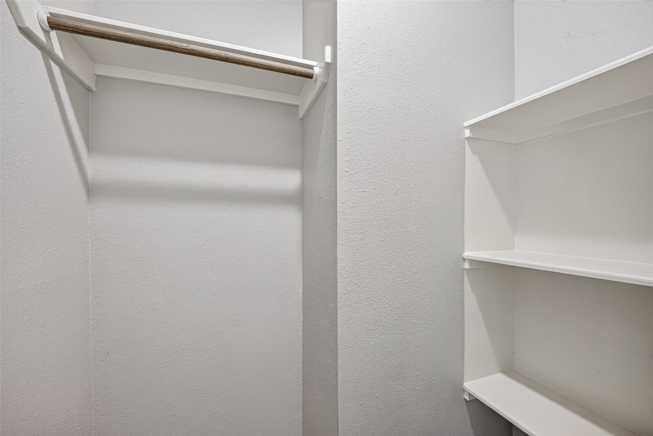 This photo shows a small walk-in closet with white walls, featuring a wooden hanging rod on the left and built-in shelving on the right, offering practical storage options.