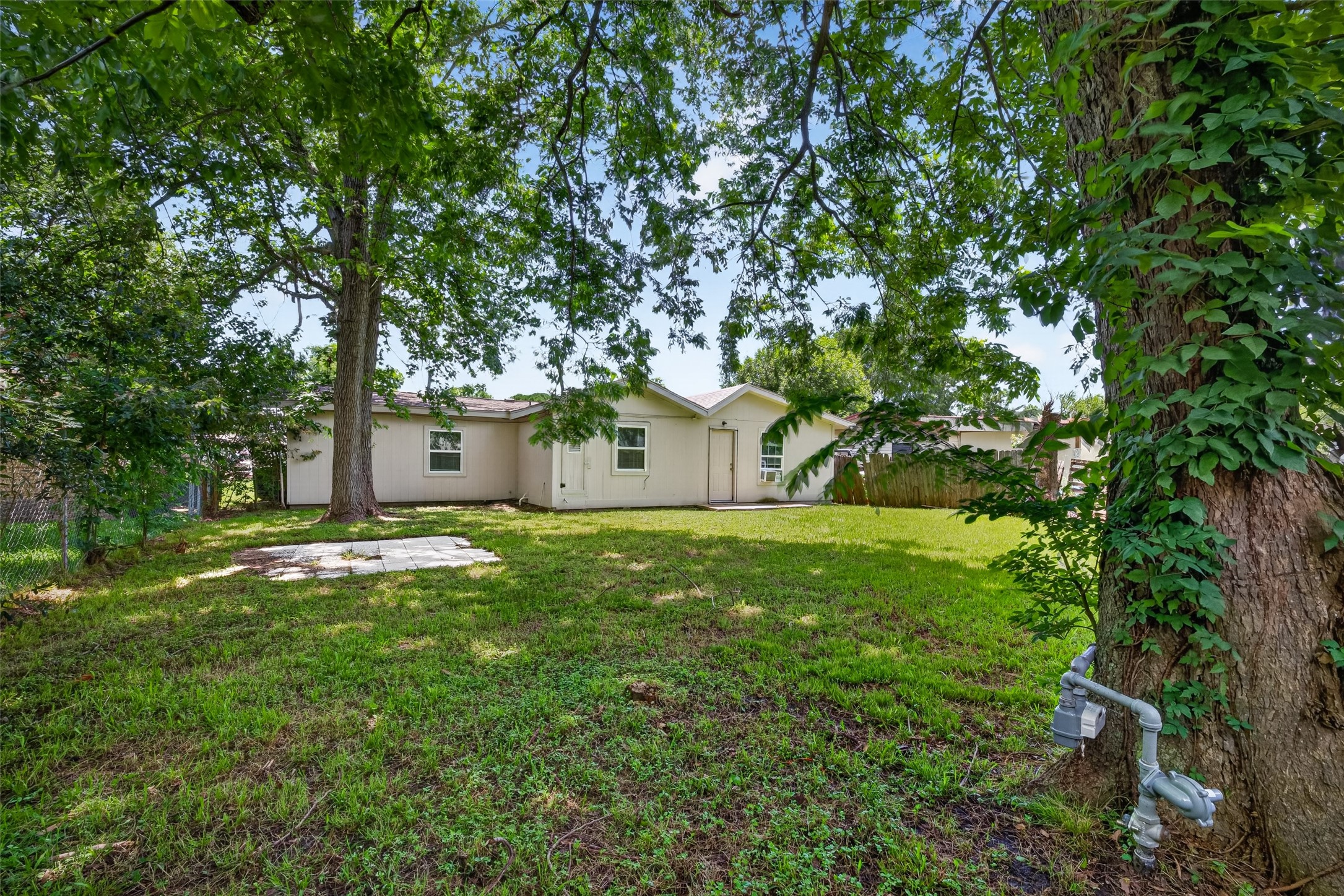 This photo shows a cozy backyard with mature trees providing shade, a grassy area, and a charming single-story home with light-colored siding. It's a peaceful, private space perfect for relaxing or entertaining.