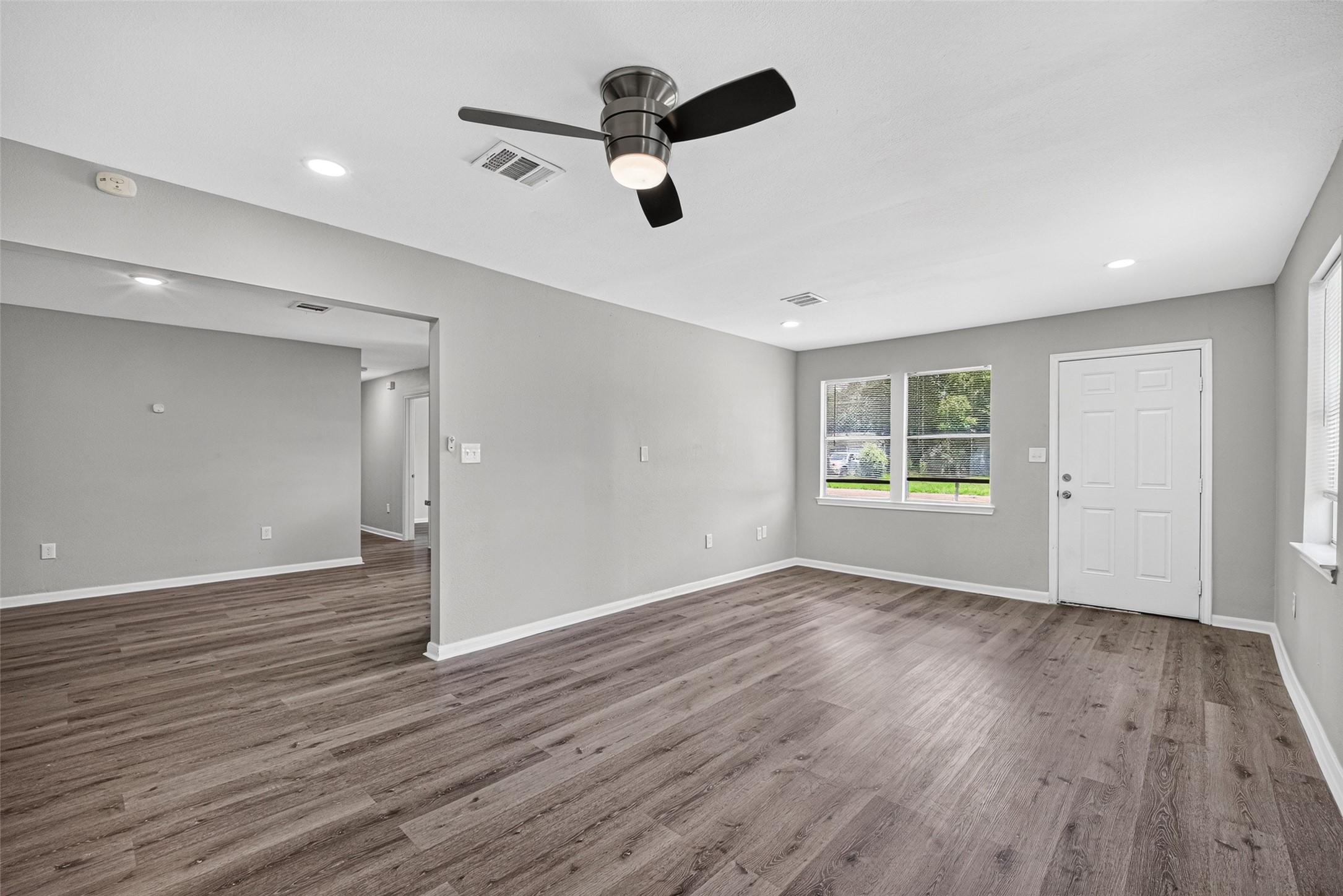 Experience a bright and open living area with sleek wood flooring and neutral gray walls. Enjoy abundant natural light from large windows, complemented by recessed lighting and a ceiling fan. This versatile space seamlessly connects to an additional area, perfect for various setups.