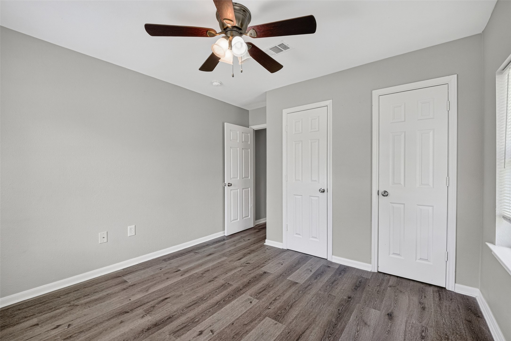 This room features modern gray walls, wood-look flooring, a ceiling fan with lights, and ample natural light from a window. It includes two white doors, likely for closet and entry access, creating a clean and functional space.