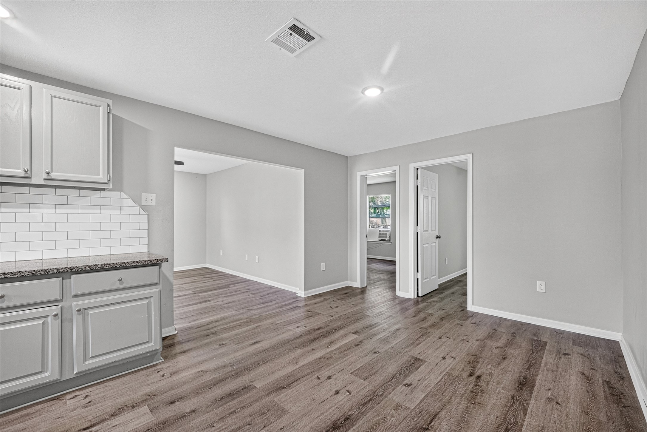 This photo shows a bright, open-concept living space with modern gray cabinets, white subway tile backsplash, and dark wood flooring. Two doorways lead to adjacent rooms, offering a spacious and versatile layout.