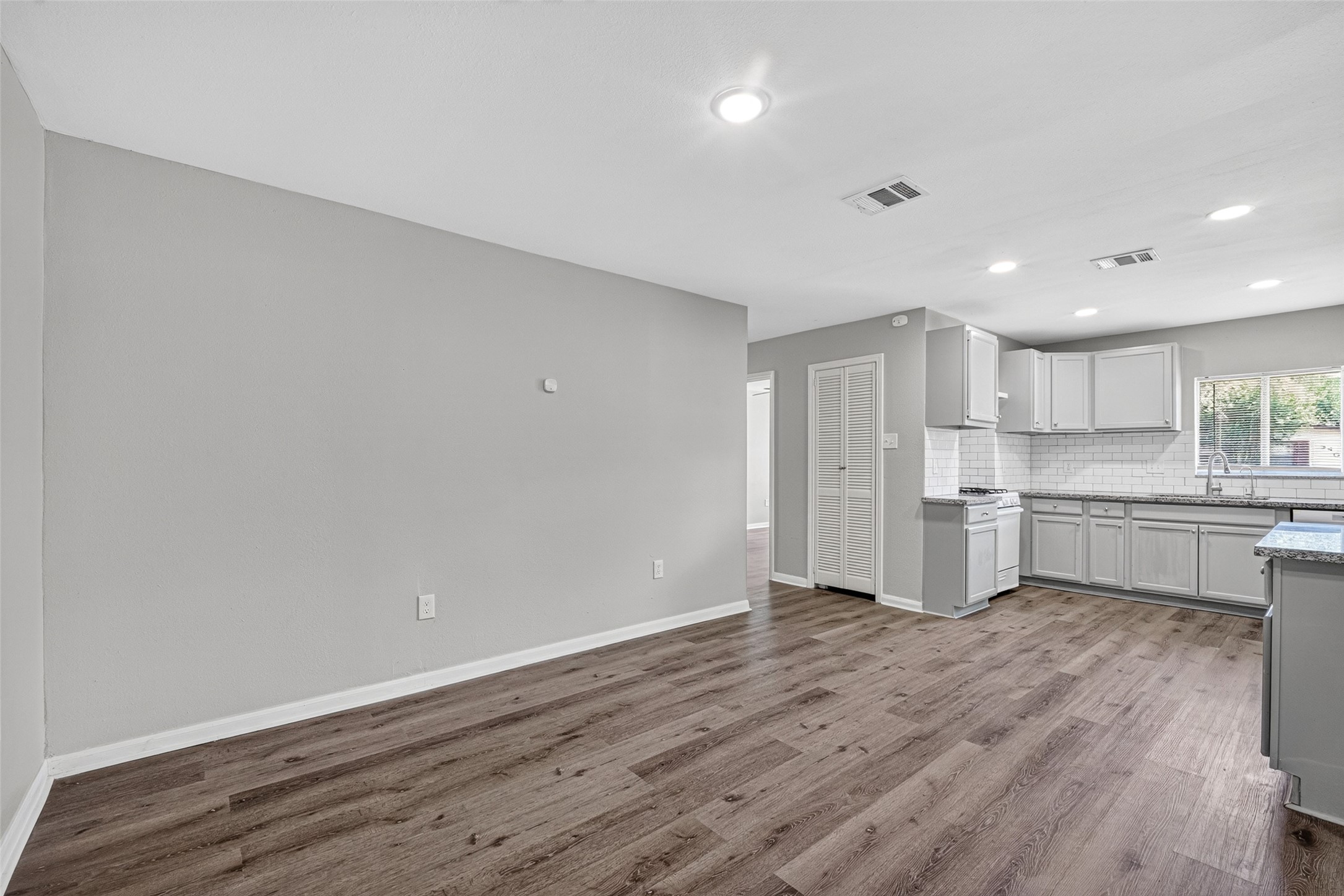 Discover a sleek, open-plan kitchen and living area with light gray walls, recessed lighting, and elegant wood-look flooring. The kitchen boasts white cabinets, a subway tile backsplash, and a window bathing the space in natural light.
