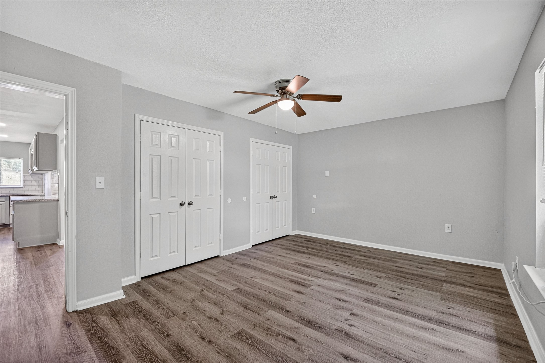 Here's a concise and compelling description for a home buyer:

"Discover this spacious room with elegant wood-look flooring and neutral gray walls. It features a ceiling fan, two double-door closets, and an open doorway leading to a bright kitchen with modern cabinetry.