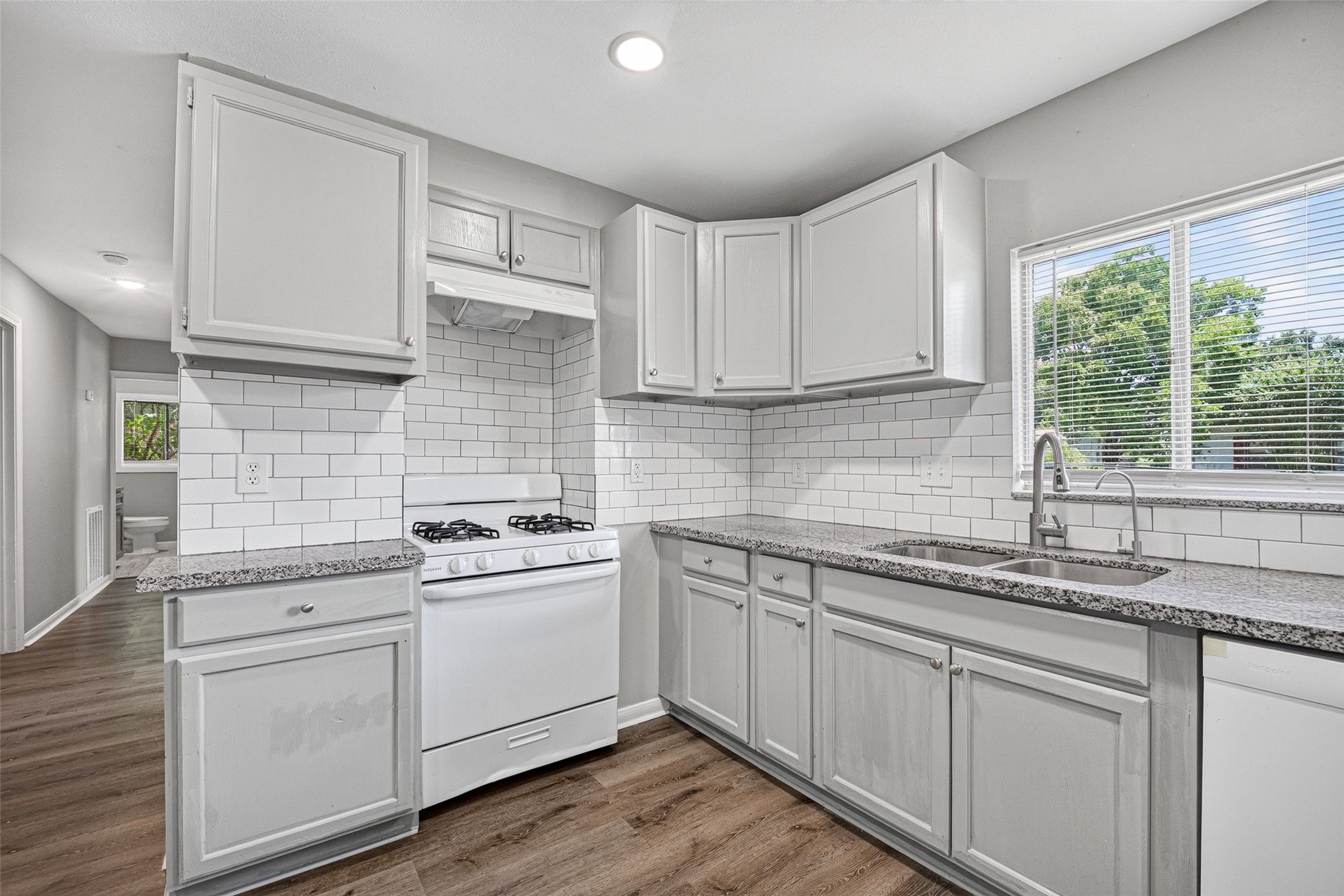 This kitchen features modern gray cabinetry, granite countertops, and a classic subway tile backsplash. It includes a gas stove and a double sink under a bright window, complemented by wood-style flooring.