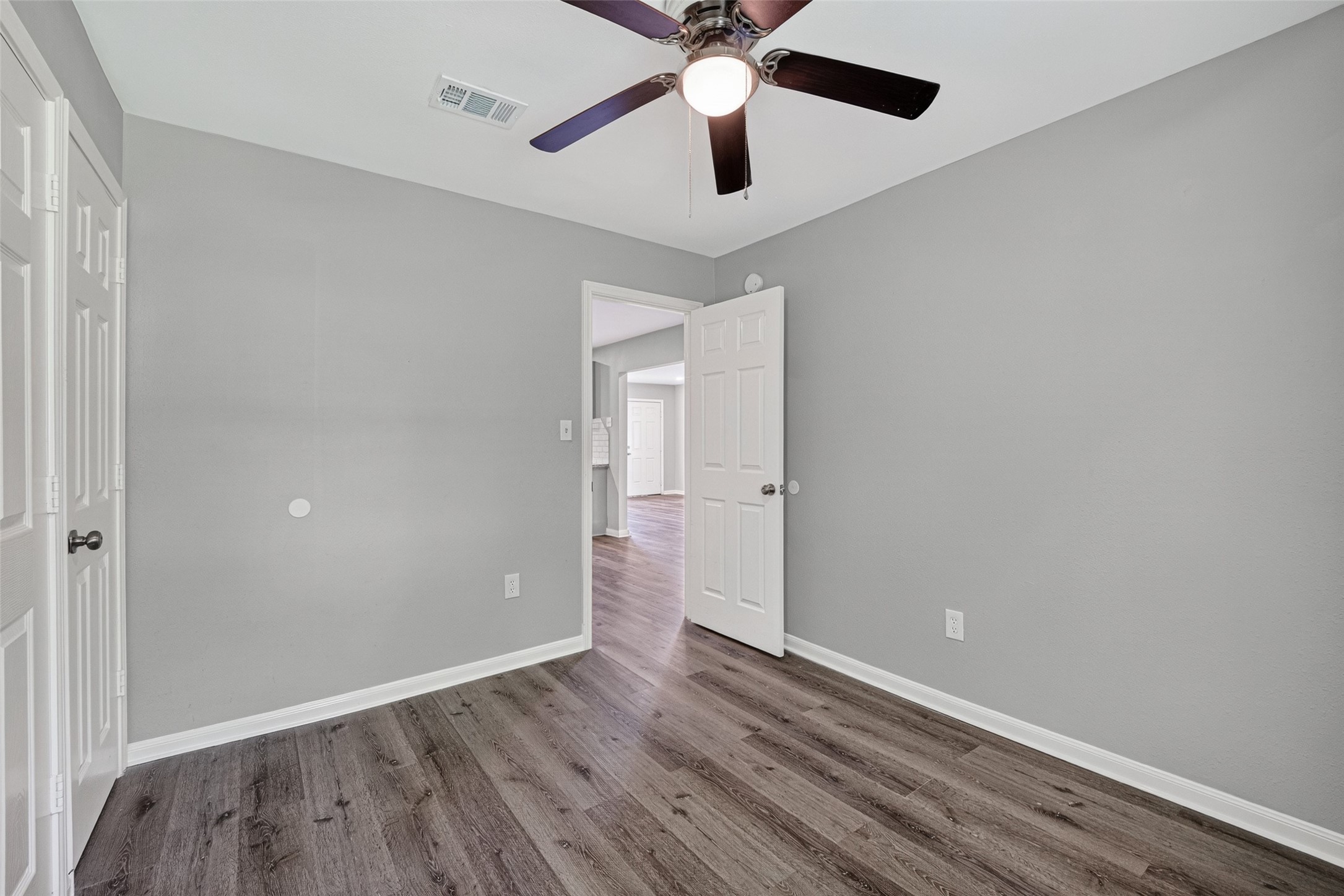 This room features gray walls, wood flooring, a ceiling fan with lights, and a white door leading to another area. It's bright and modern, perfect for a bedroom or office space.