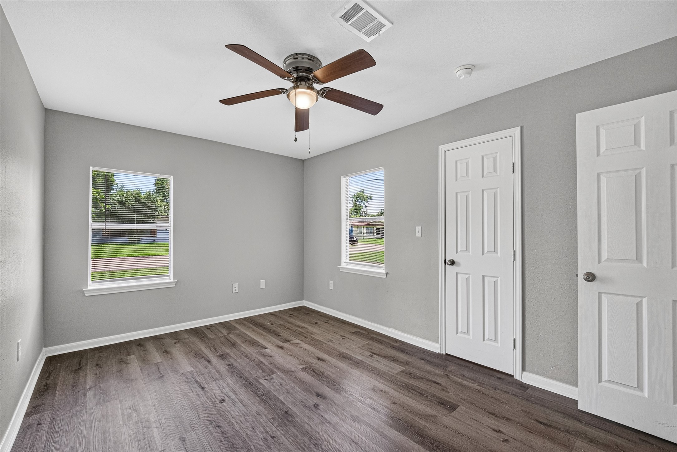 This room features a modern, neutral design with gray walls and dark wood flooring. It has two windows for natural light, a ceiling fan, and a closet with white doors.