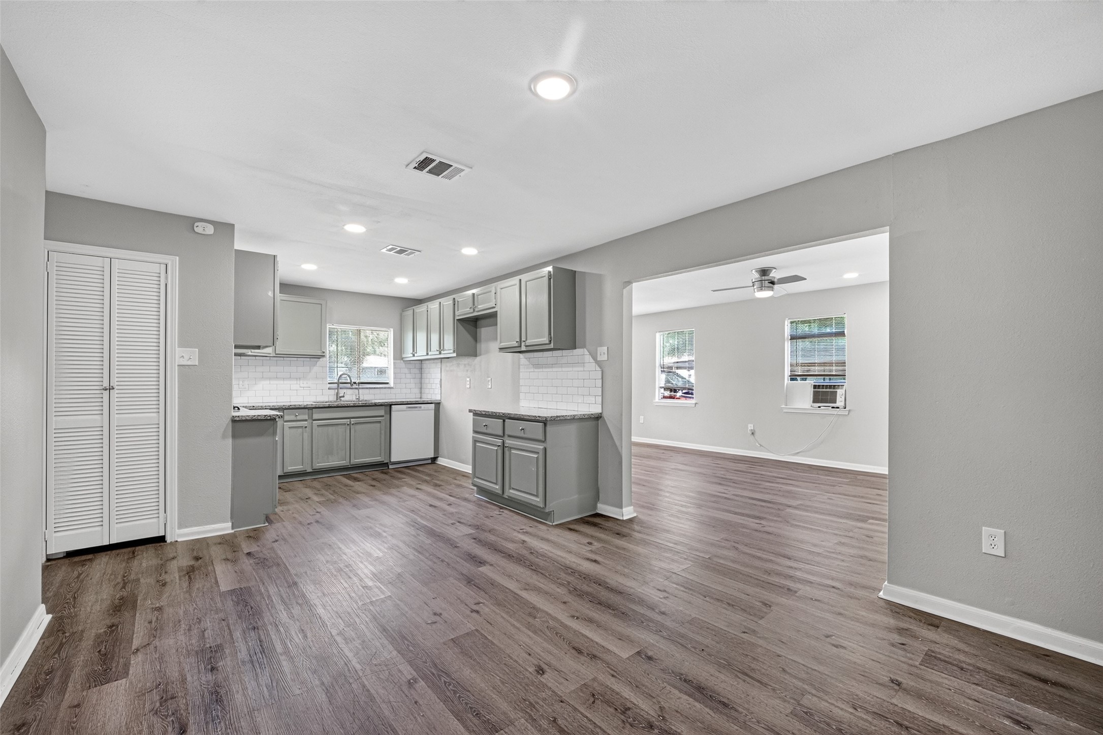 Experience modern living in this open-concept kitchen and living area, featuring sleek gray cabinetry, elegant hardwood flooring, and abundant natural light. The kitchen boasts a stylish white tile backsplash and recessed lighting, seamlessly flowing into a bright living space with large windows and a ceiling fan.