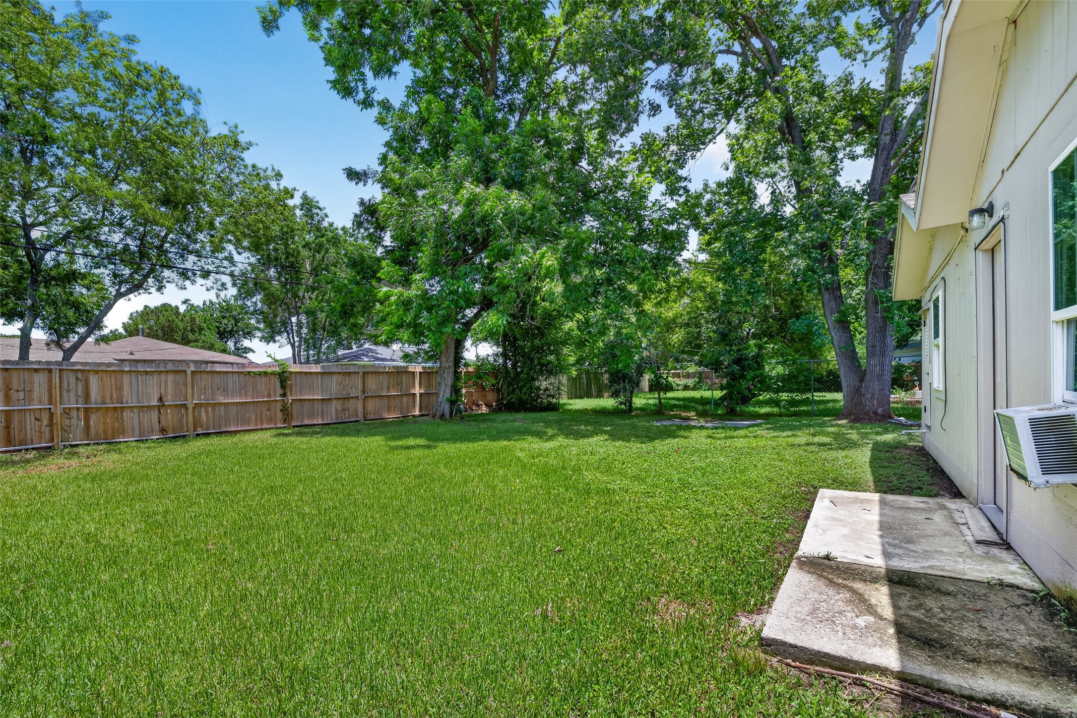 This photo shows a spacious backyard with lush green grass and mature trees, enclosed by a wooden privacy fence. The side of the house, with a window and exterior door, is partially visible. Ideal for outdoor activities and relaxation.