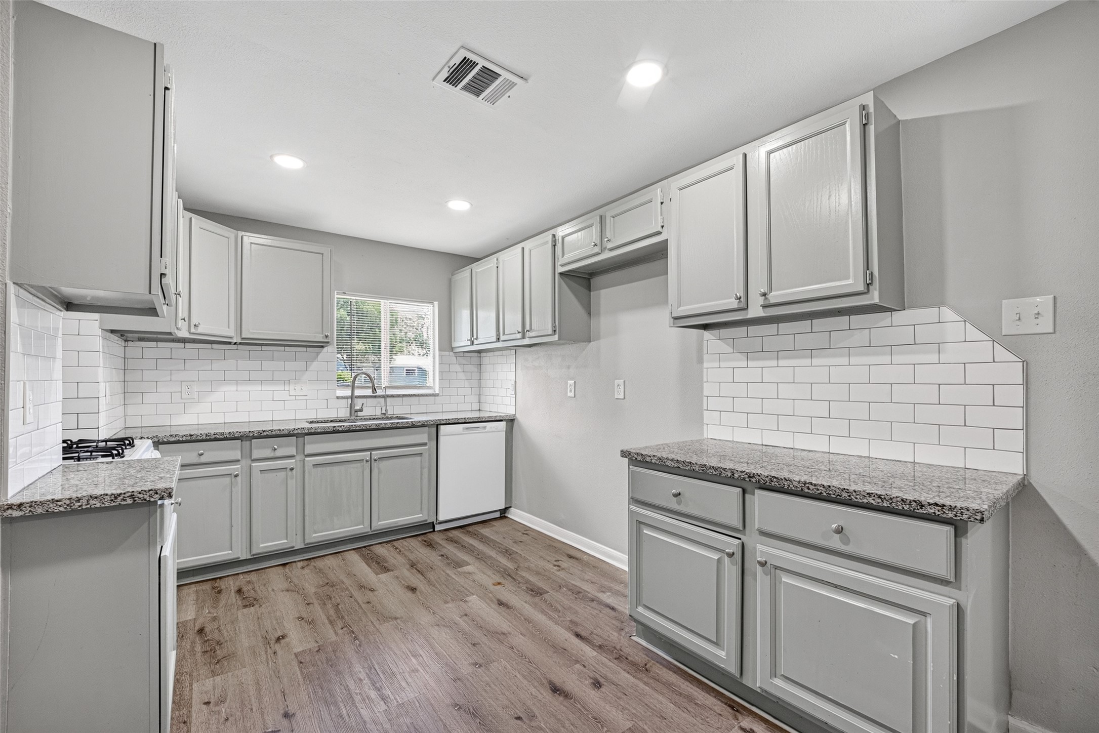 This kitchen features modern gray cabinets, granite countertops, and a white subway tile backsplash. It has a gas cooktop, dishwasher, and wood-look flooring, creating a stylish and functional space.