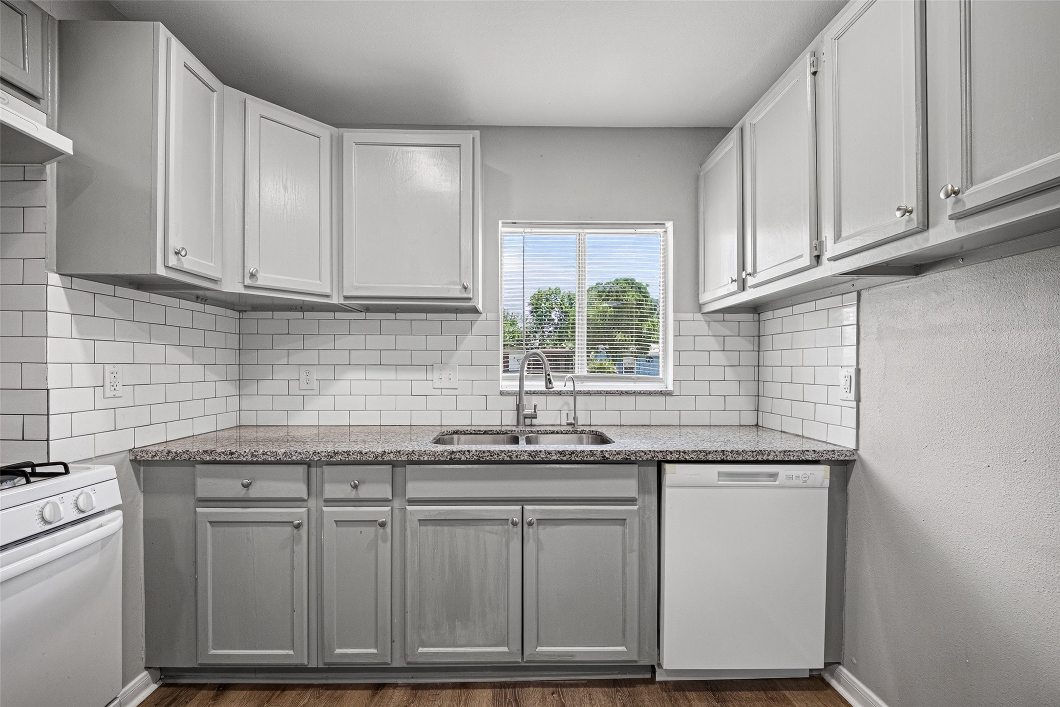 This kitchen features modern, light gray cabinets with sleek hardware, a granite countertop, and a subway tile backsplash. It includes a window above the sink for natural light, a dishwasher, and a gas stove, all complemented by wood-style flooring.
