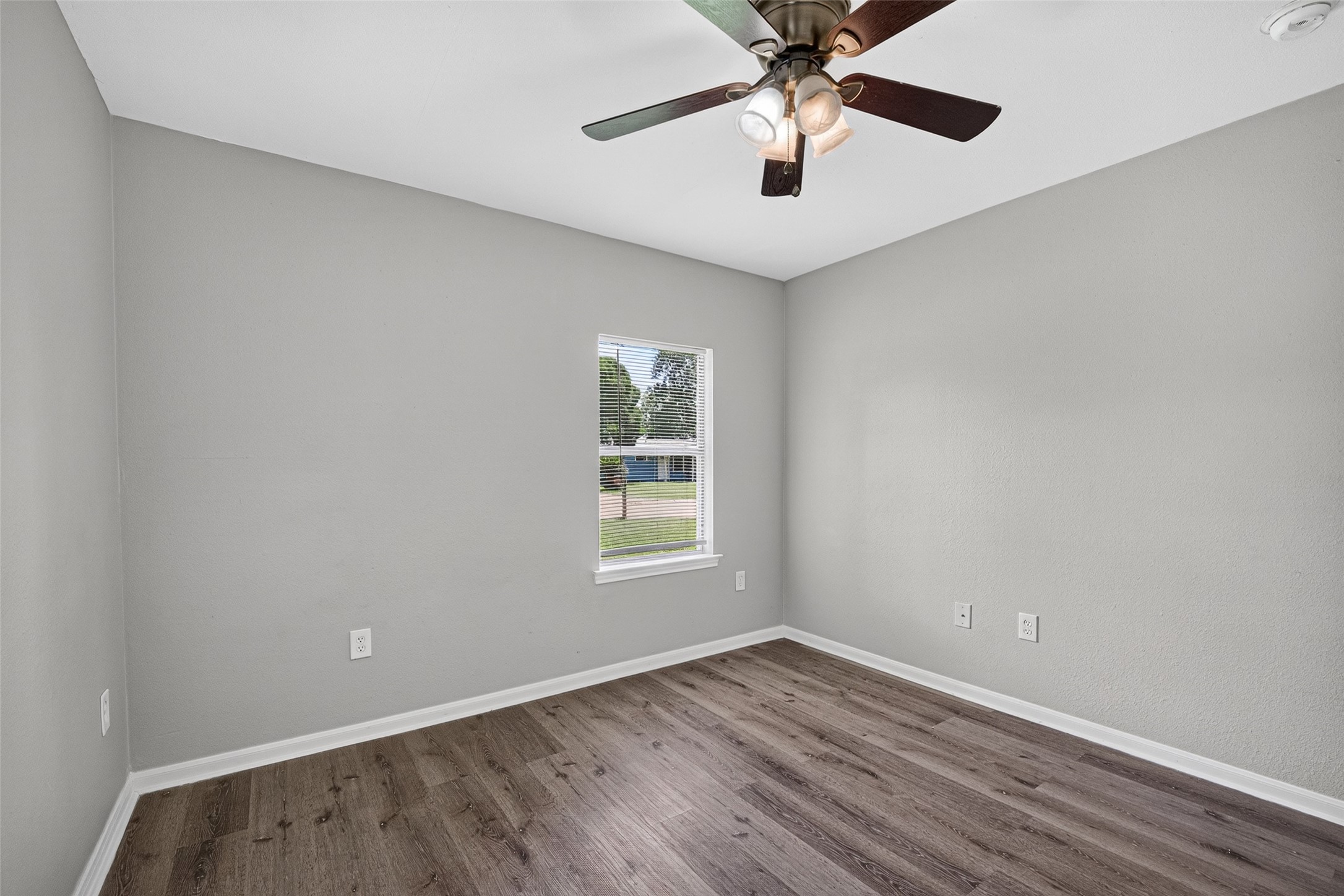 This is a bright, unfurnished room with light gray walls, dark wood flooring, and a ceiling fan. A window provides natural light and a view of the outside.
