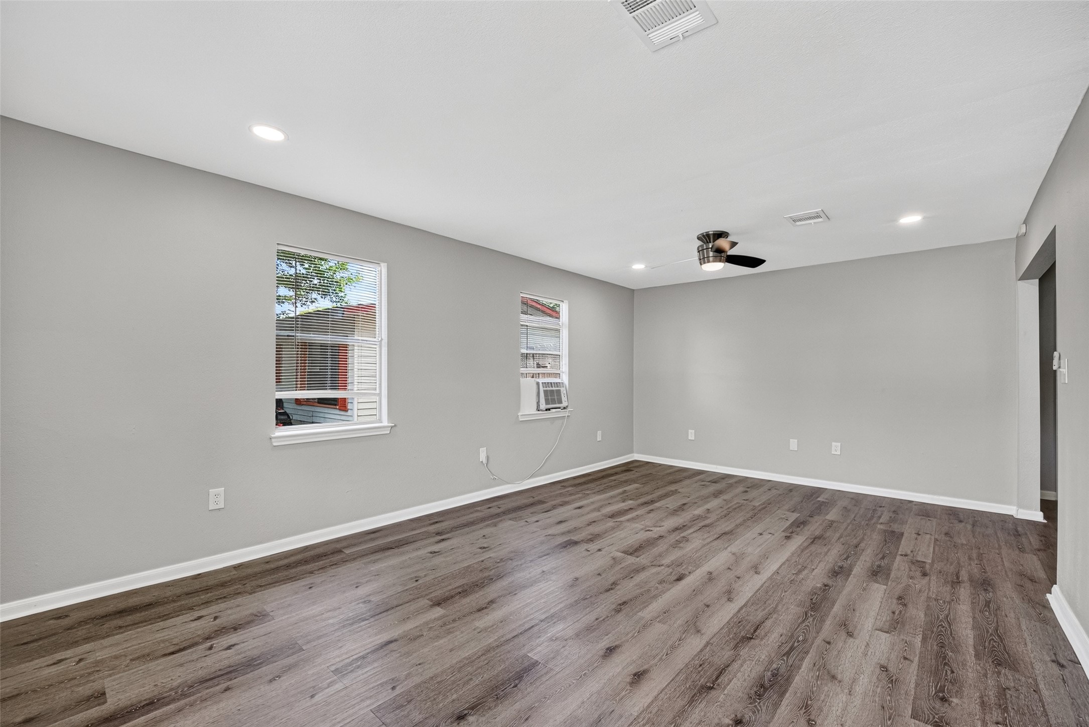 Spacious living room with sleek hardwood floors, neutral gray walls, and recessed lighting. Features two windows for natural light and a modern ceiling fan. Ideal for comfortable living and entertaining.