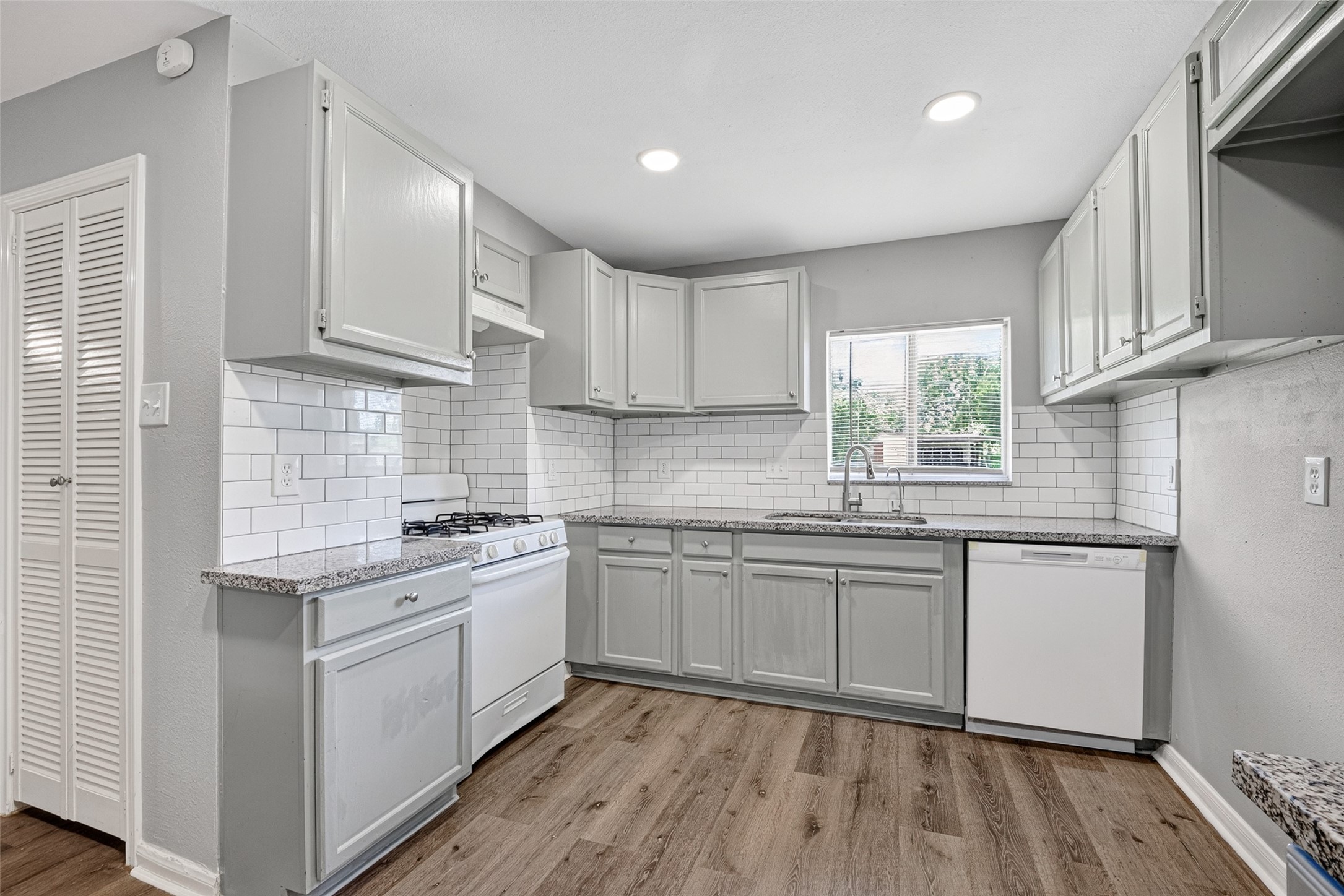 This kitchen features a modern, clean design with light gray cabinets, granite countertops, and white subway tile backsplash. It includes a gas stove, dishwasher, and a window above the sink for natural light. The space is complemented by wood-style flooring and recessed lighting.