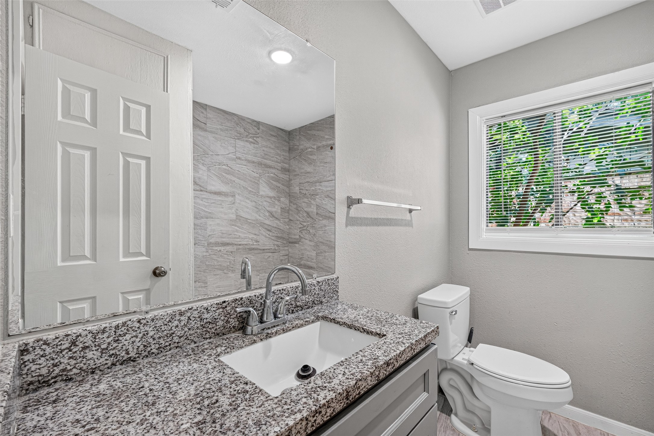 This bathroom features a sleek granite countertop, modern fixtures, and a large mirror. The space is well-lit with natural light from a window with blinds, and it has a neutral color palette for a clean and fresh look.
