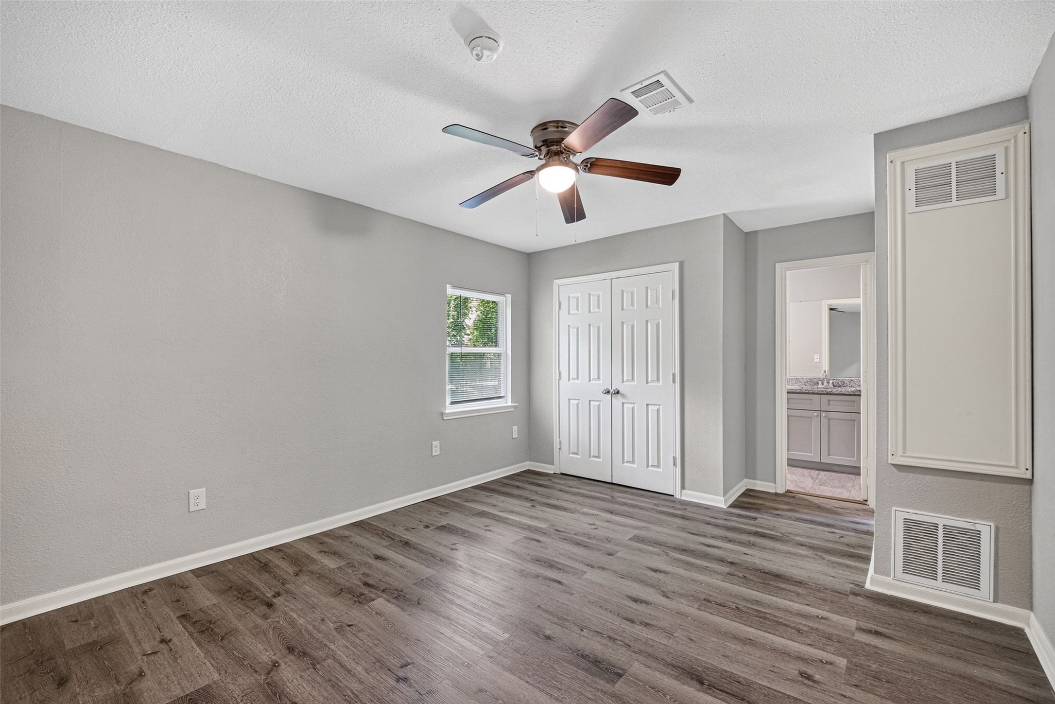 This room features fresh gray walls, wood-look flooring, and a ceiling fan. It has a window for natural light, a double-door closet, and an adjoining bathroom.