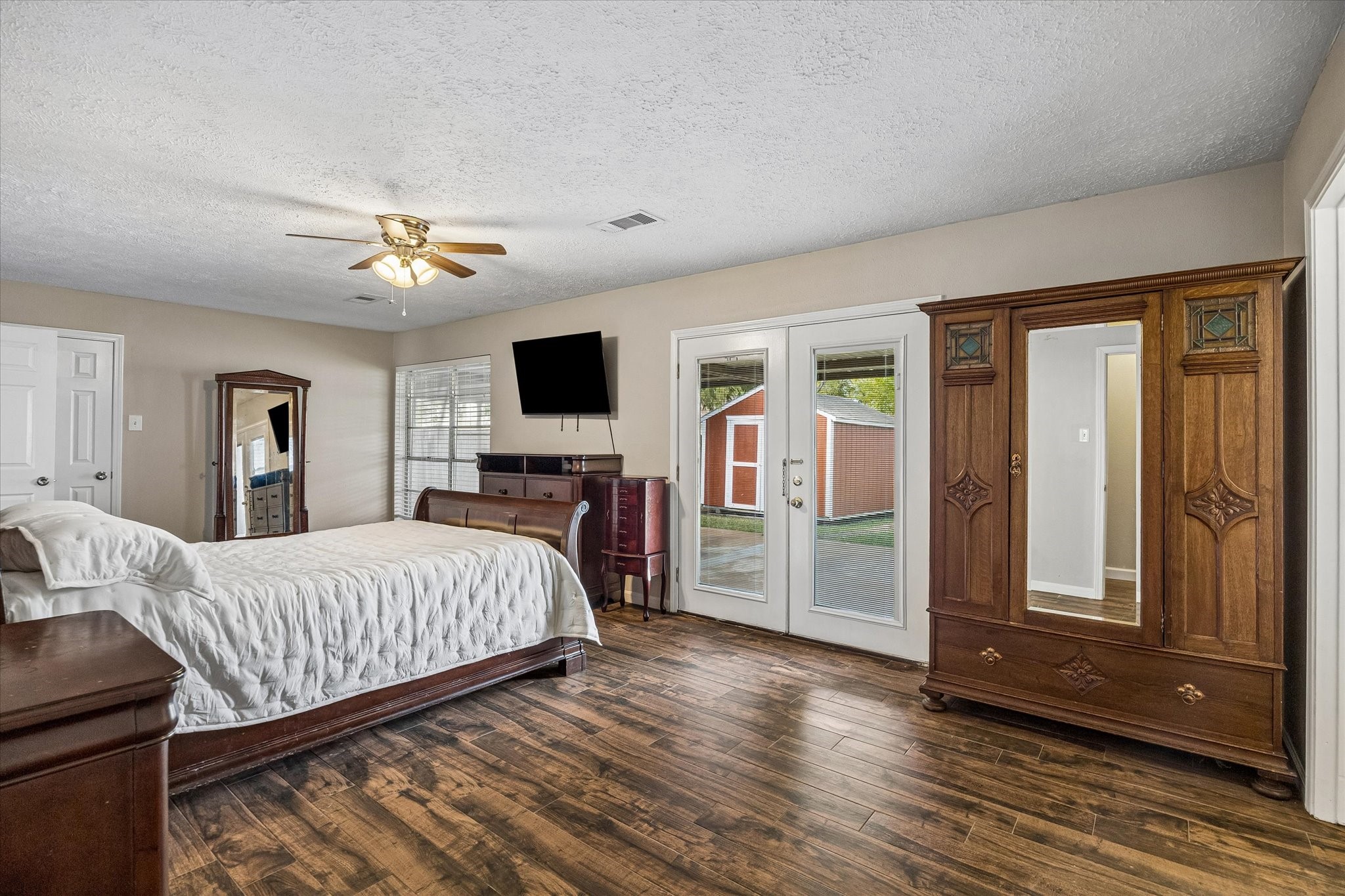 The large primary bedroom features elegant wood-look tile flooring, abundant natural light, and French doors that open to a peaceful outdoor view, creating a bright and inviting retreat.