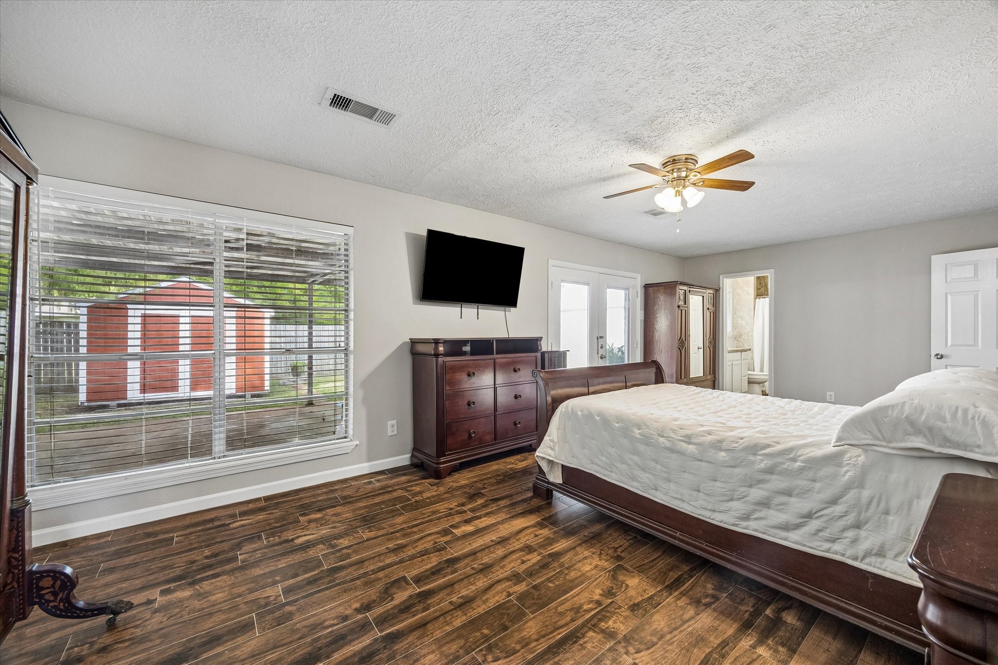 Spacious bedroom with rich wood flooring, a ceiling fan, and abundant natural light from large windows overlooking the backyard.