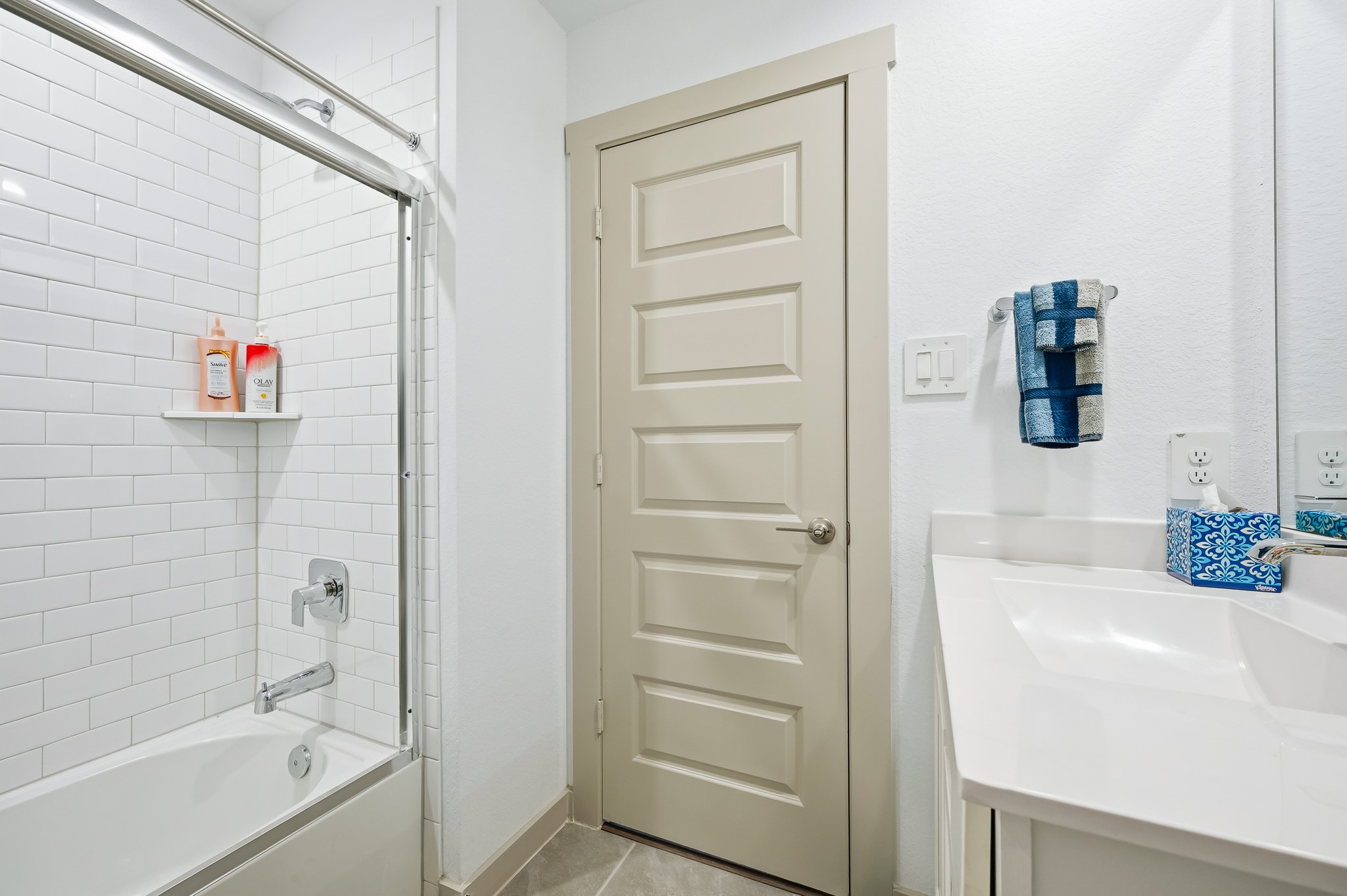 This bathroom features a clean, modern design with white subway tiles in the shower, a sleek white vanity, and a neutral-toned door. The space is bright and functional, with convenient shelving and a towel rack.