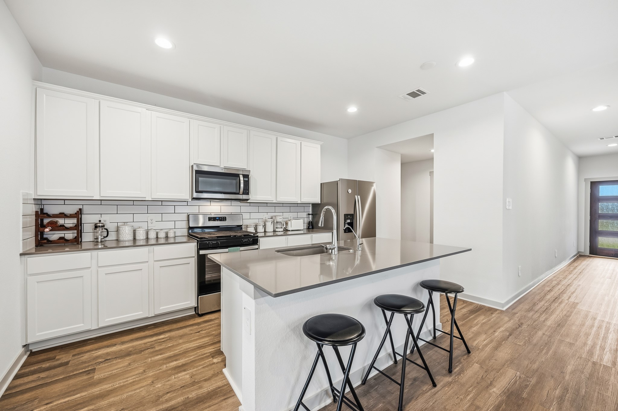 This modern kitchen features sleek white cabinetry, stainless steel appliances, and a subway tile backsplash. It includes a spacious island with a sink and bar seating, complemented by wood-look flooring and recessed lighting for a bright, inviting atmosphere.