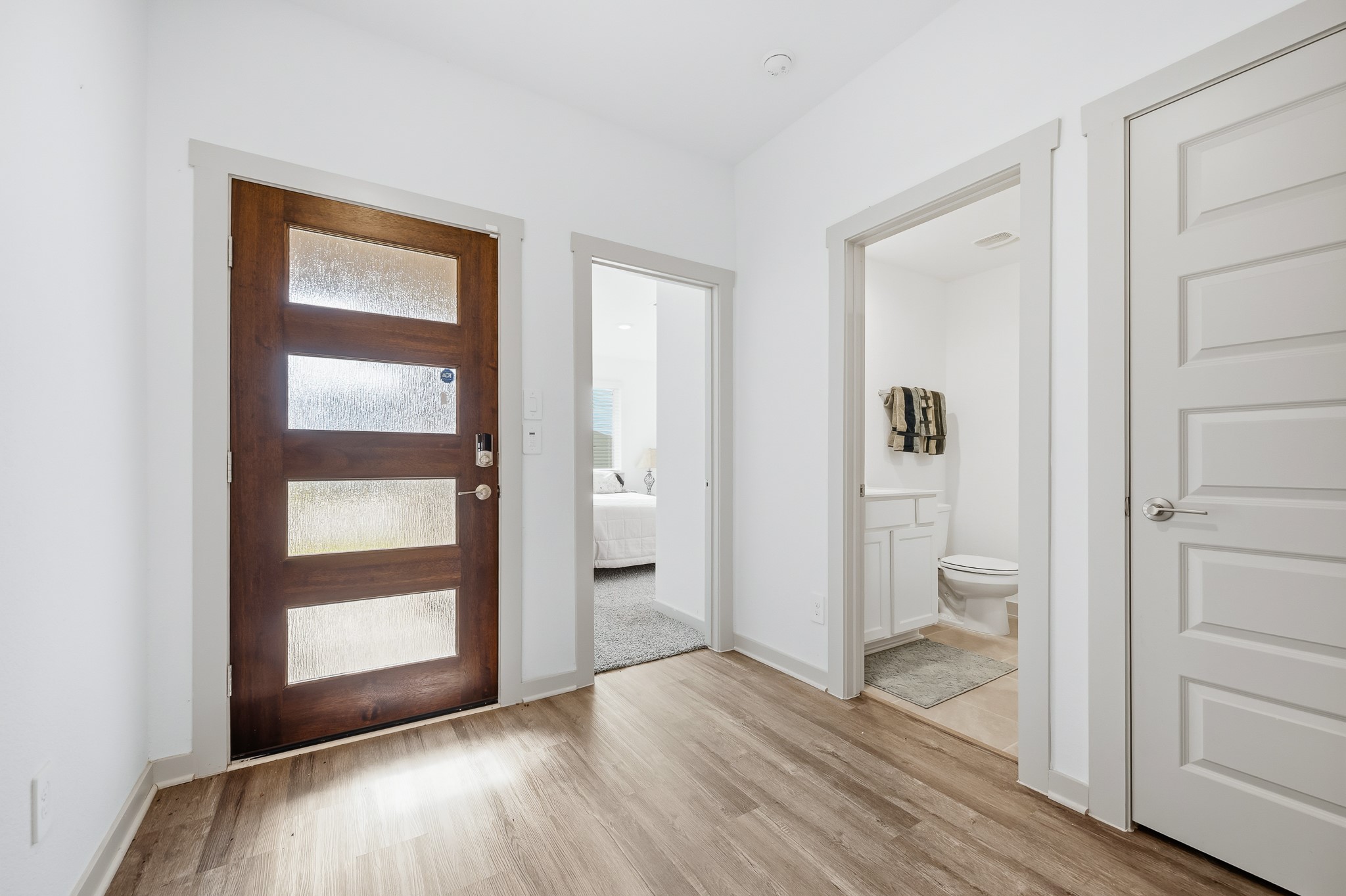 Bright entryway featuring a modern wooden front door with glass panels. The space connects to a carpeted bedroom and a bathroom with a white vanity. Stylish and functional layout.