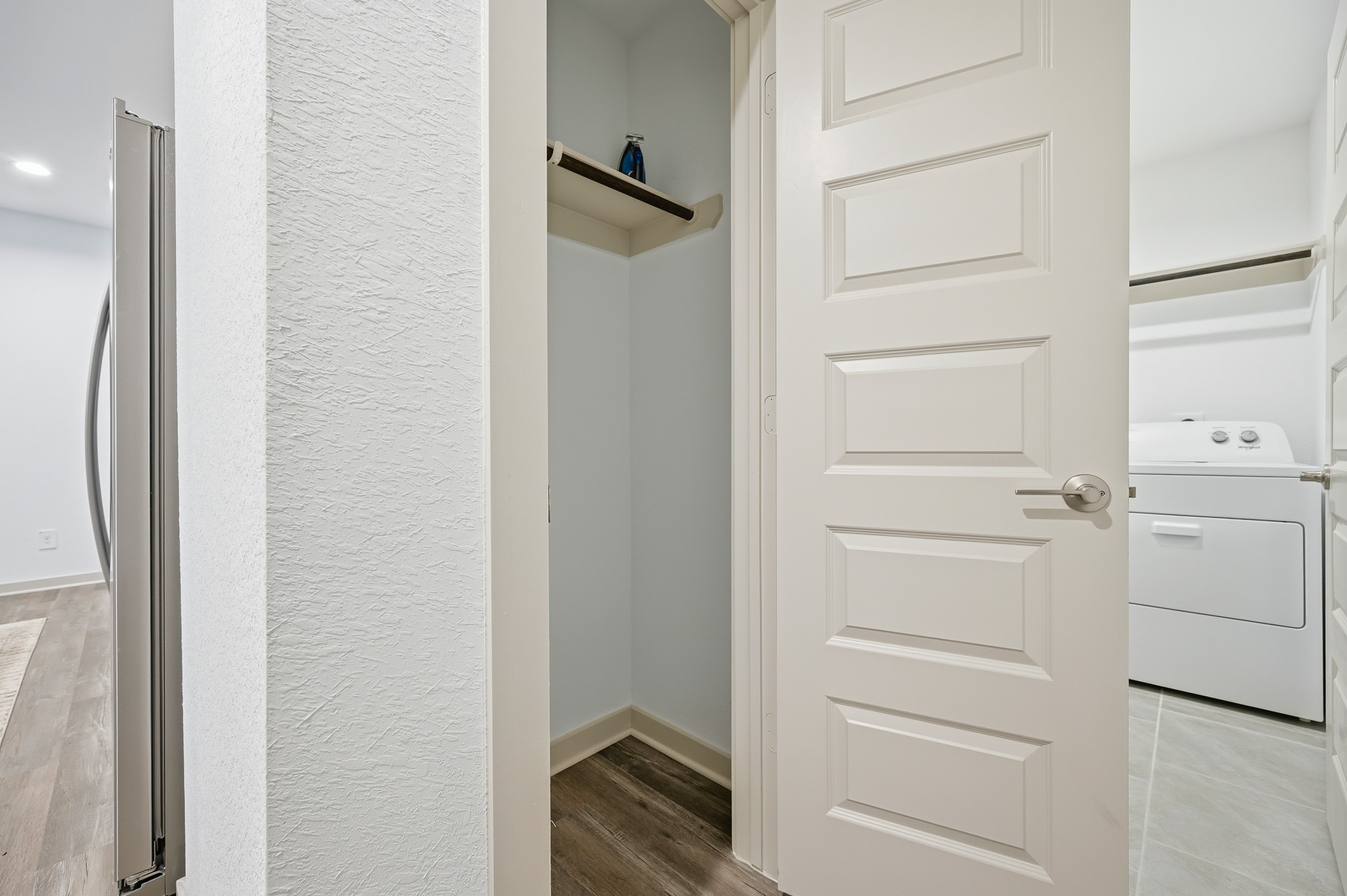 This photo showcases a small interior space with a modern white door and a closet with shelving. Adjacent is a laundry area featuring a washer and dryer, set on a tiled floor. The area is bright with light walls and wood-style flooring.