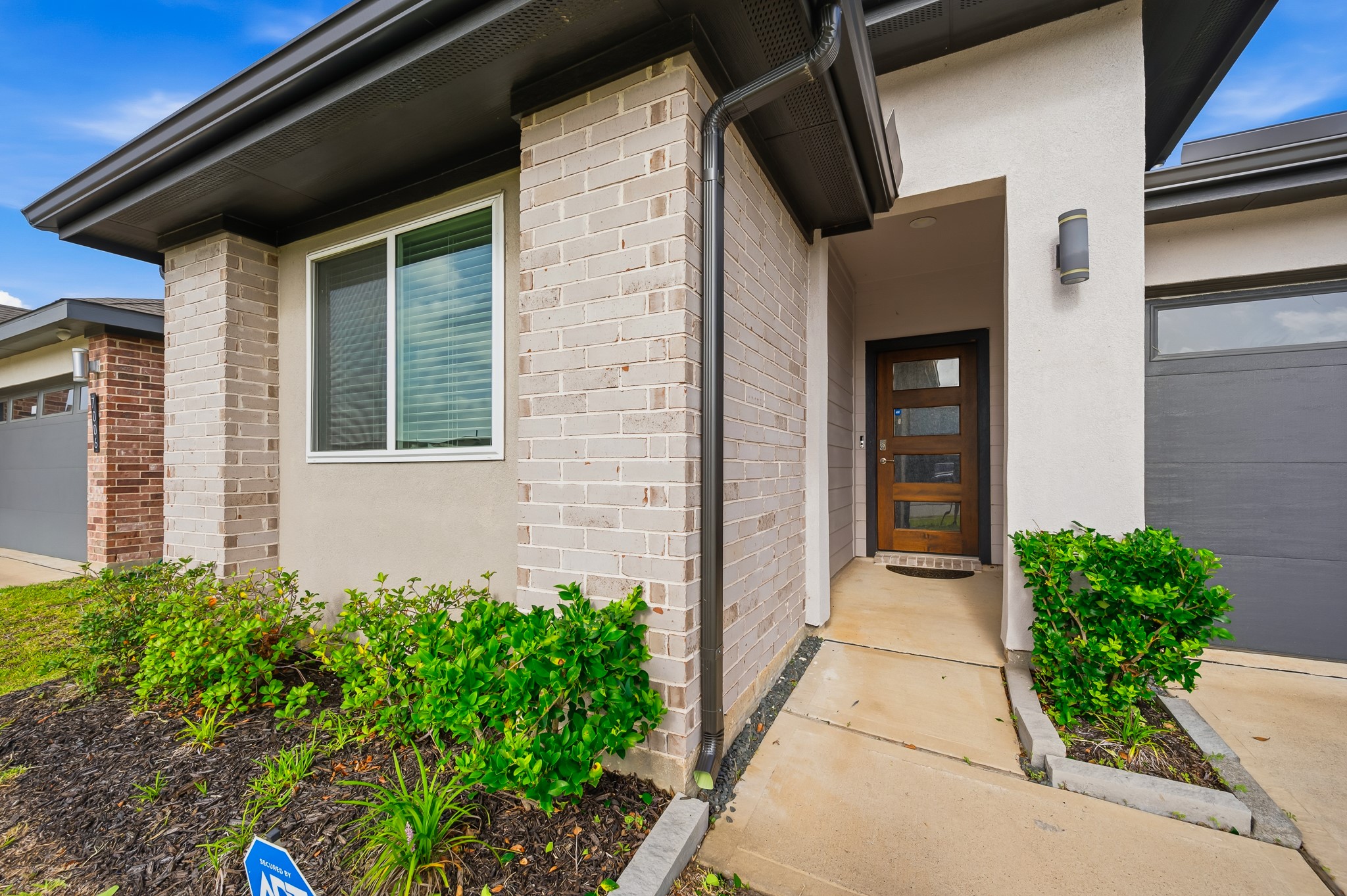 This modern home features a welcoming entrance with a wooden door, contemporary light fixture, and attractive brick accents. The well-maintained landscaping adds to its curb appeal.