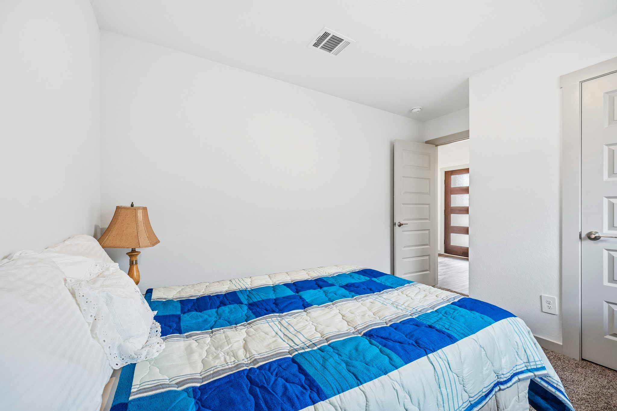 This cozy bedroom features a modern design with white walls, a blue and white quilt on the bed, and a bedside lamp. The room is well-lit and has an open door leading to a hallway, enhancing the sense of space.