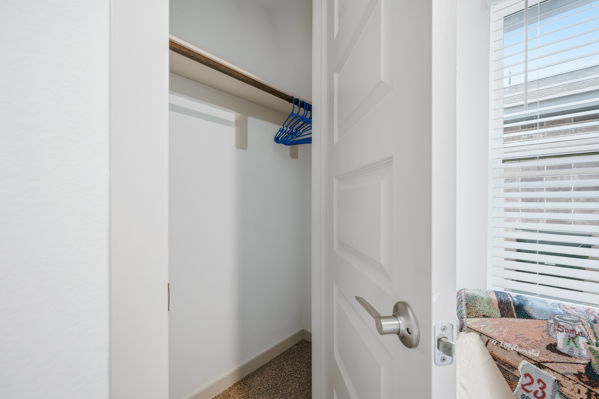 A small, white closet with a wooden rod and blue hangers, carpeted flooring, and an open door. Nearby, there's a window with blinds allowing natural light to enter.