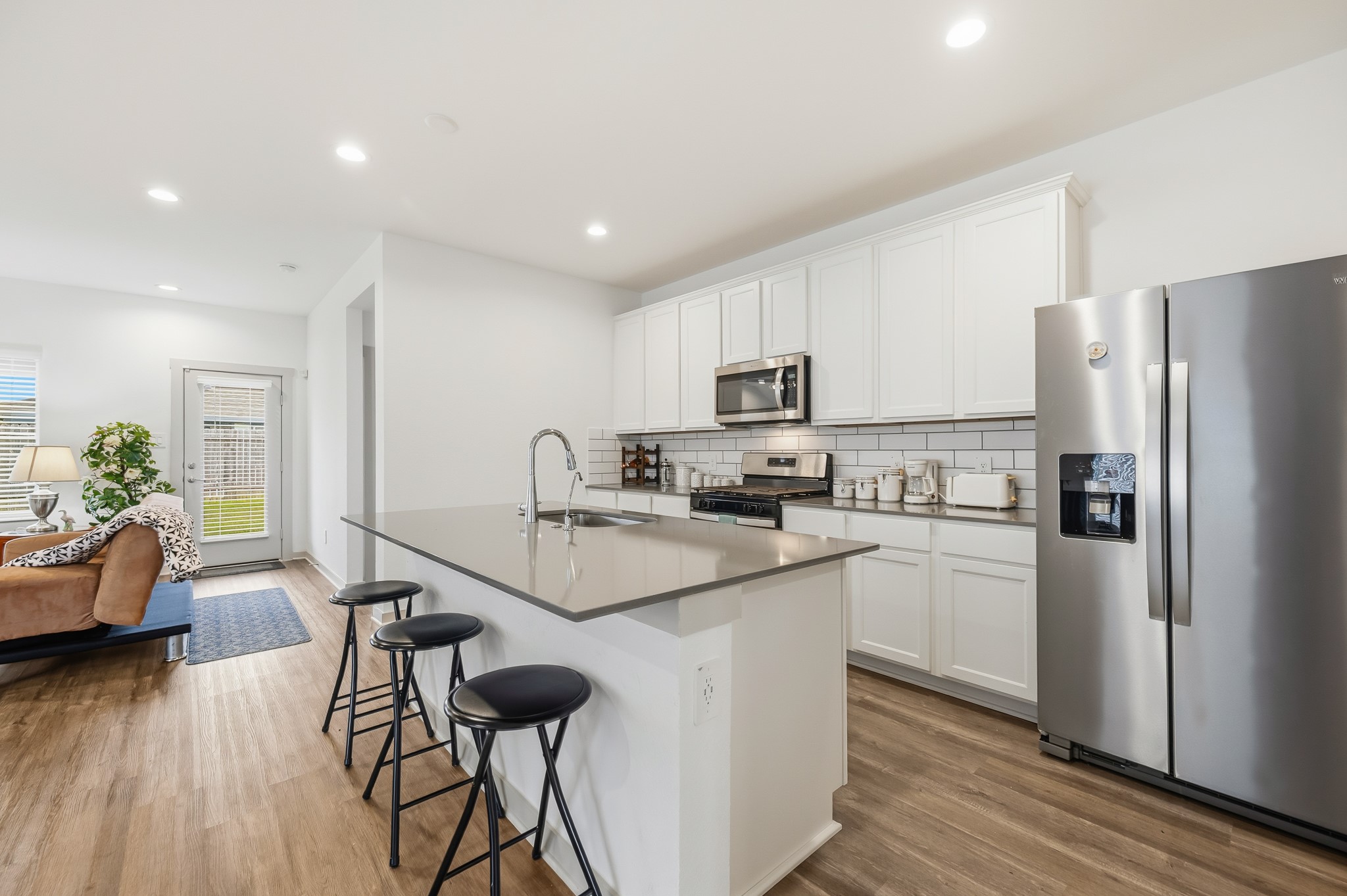 This modern kitchen features sleek white cabinetry, stainless steel appliances, and a spacious island with bar seating. It opens into a cozy living area with hardwood floors and natural light from a glass door leading outside.