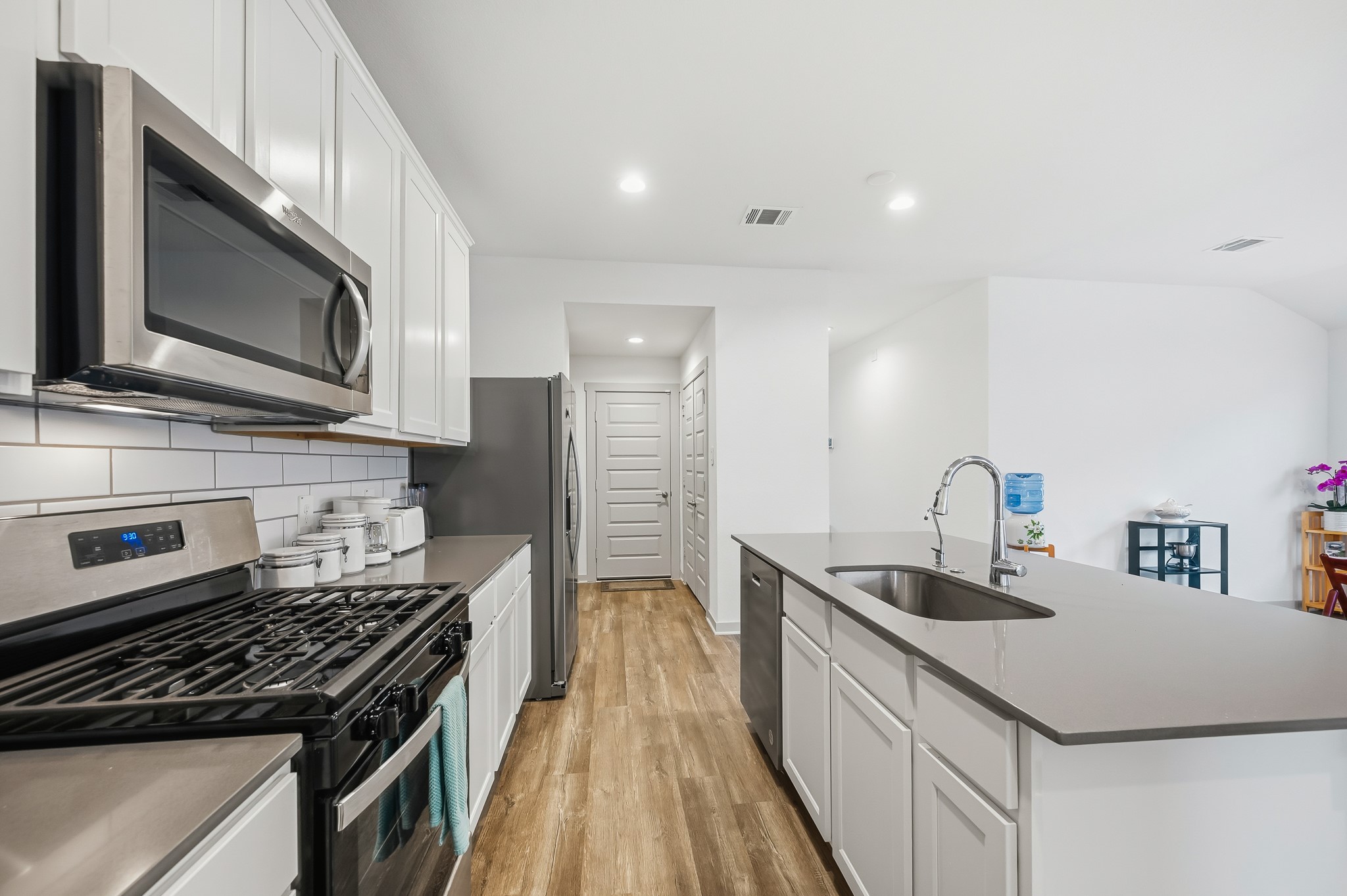 Bright and modern kitchen featuring stainless steel appliances, sleek countertops, white cabinetry, and a spacious island with a sink. The open layout is enhanced by recessed lighting and wood-style flooring, providing a welcoming space for cooking and entertaining.