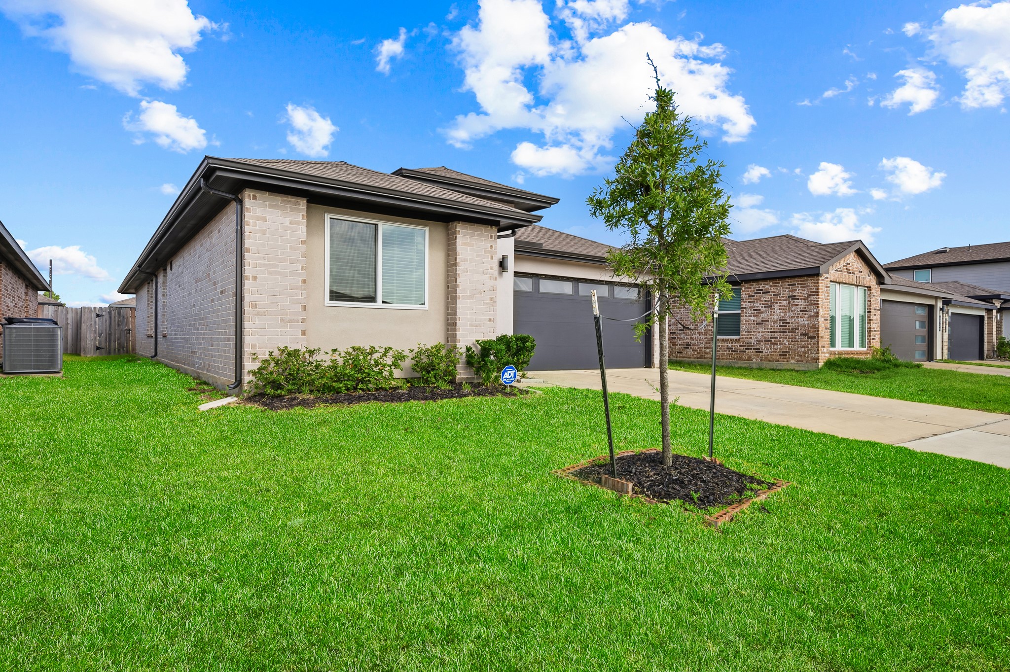 This photo shows a modern, single-story brick home with a well-maintained lawn and a young tree in the front yard. The house has a light brick exterior, large windows, and a two-car garage. It's situated in a suburban neighborhood with similar style homes.