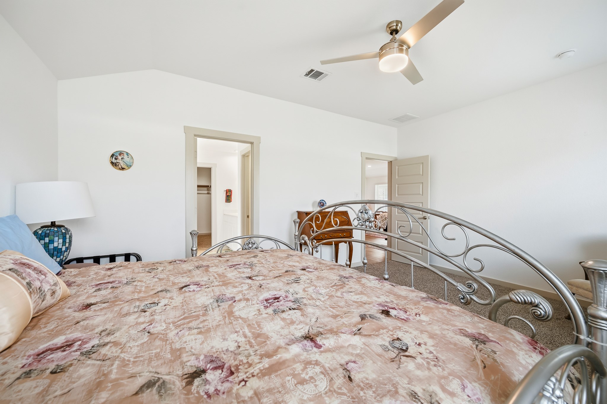 This bright bedroom features a decorative metal bedframe, floral bedding, and a ceiling fan. The room has neutral walls, a carpeted floor, and access to a walk-in closet.