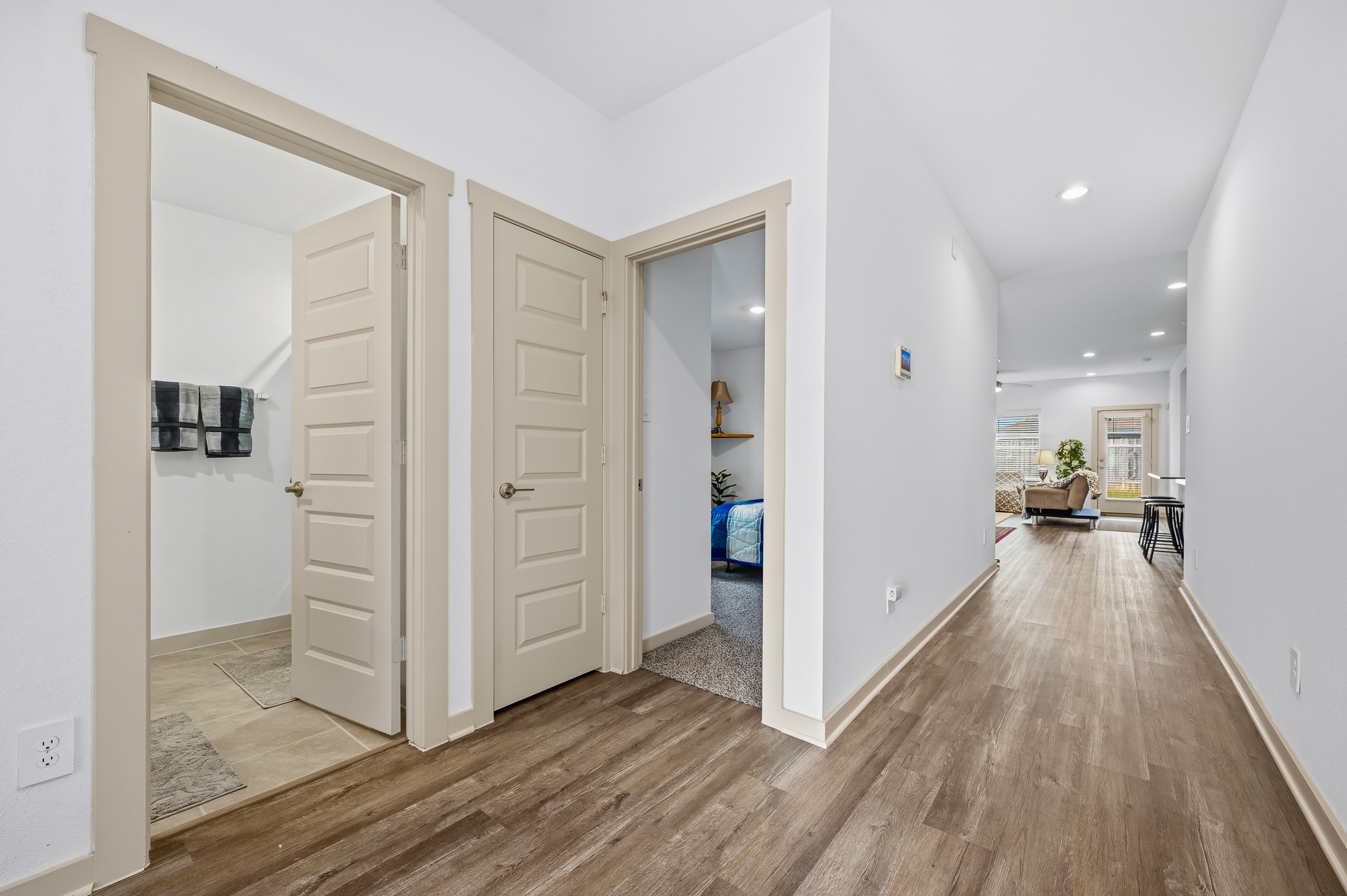 This photo showcases a modern hallway with wood-style flooring, leading to a bright living area. It features a bathroom on the left and a bedroom on the right, both with beige door frames. The space is well-lit with recessed lighting.