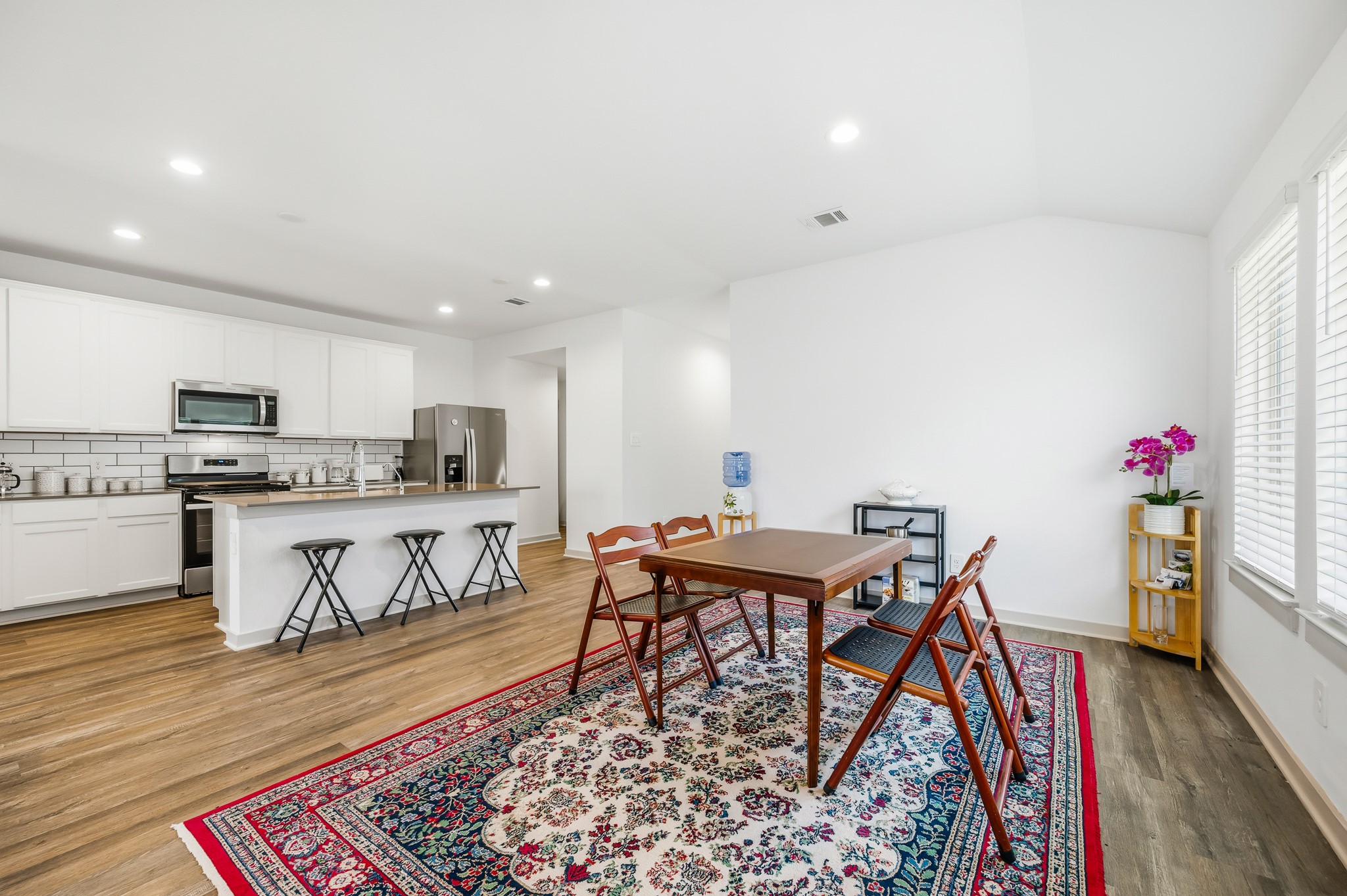 This is a bright, open-concept kitchen and dining area with modern white cabinetry, stainless steel appliances, and a spacious island with bar stools. The dining area features a warm wood floor and a colorful area rug, creating an inviting space for meals and gatherings.