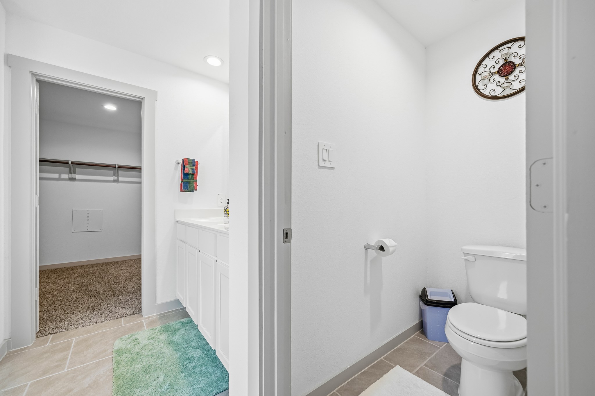 This photo shows a bright bathroom with a separate toilet area, featuring white cabinetry, a colorful towel, and light tile flooring. It leads to a spacious walk-in closet with carpeted flooring.