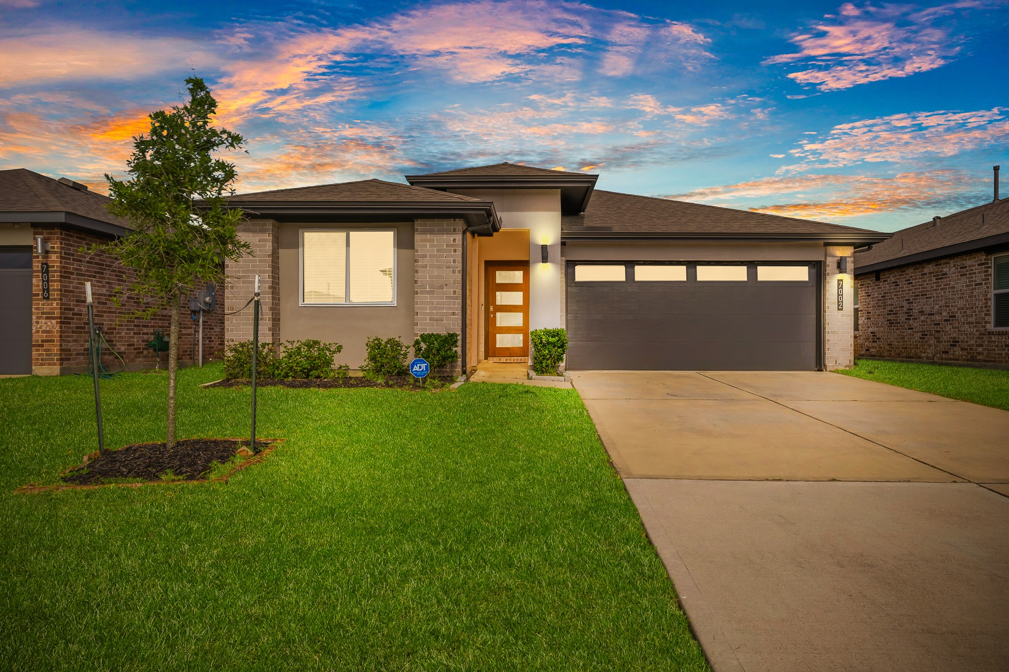 This is a modern, single-story home with brick exterior, featuring a neatly landscaped front yard, a young tree, and a spacious driveway leading to a double garage. The sunset sky adds a warm ambiance. This is a modern, single-story home with brick exterior, featuring a neatly landscaped front yard, a young tree, and a spacious driveway leading to a double garage. The sunset sky adds a warm ambiance.