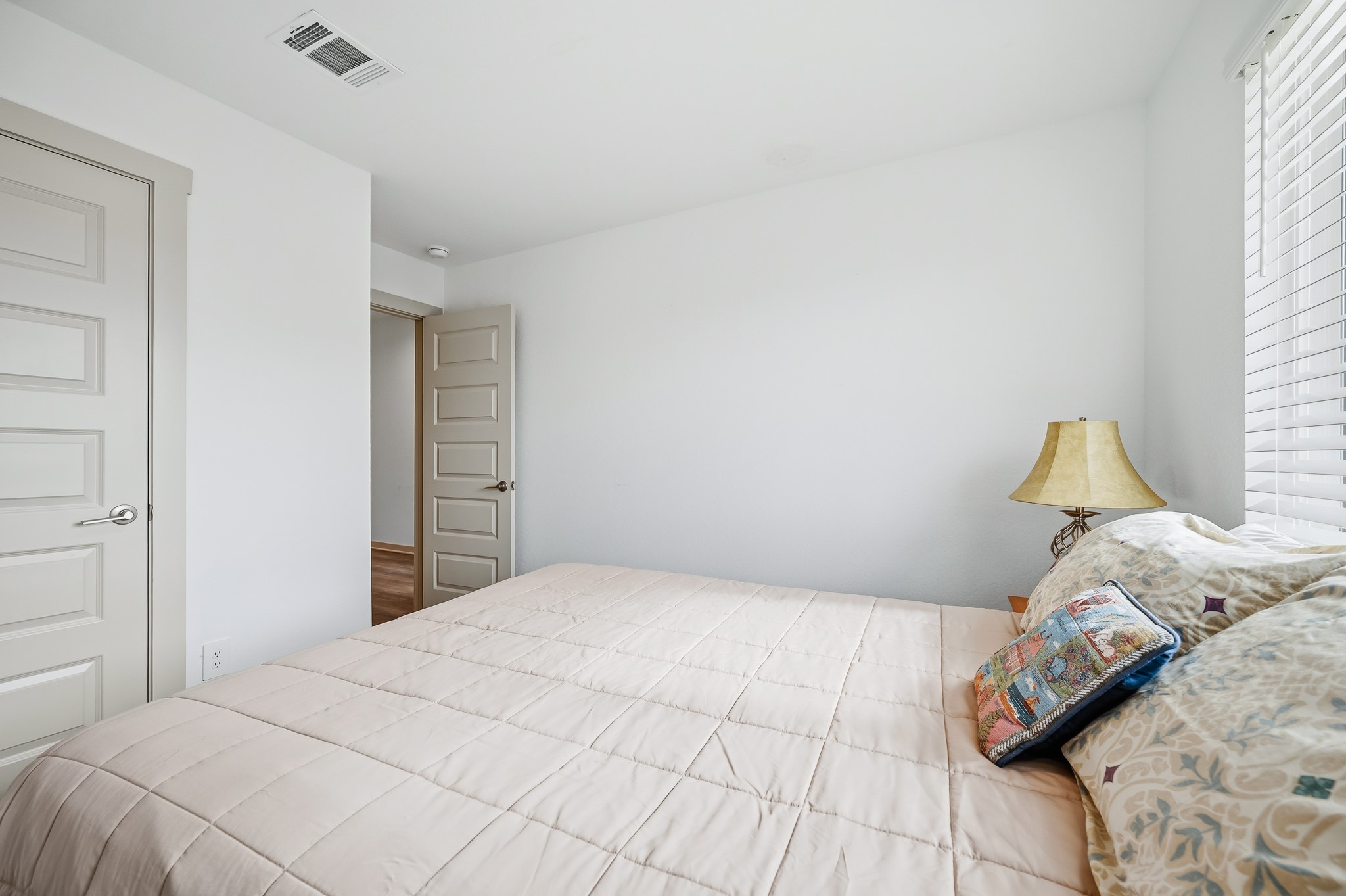 This bedroom features a neutral color scheme with ample natural light from a window. It includes a cozy bed, decorative pillows, and a bedside lamp, creating a warm and inviting atmosphere.