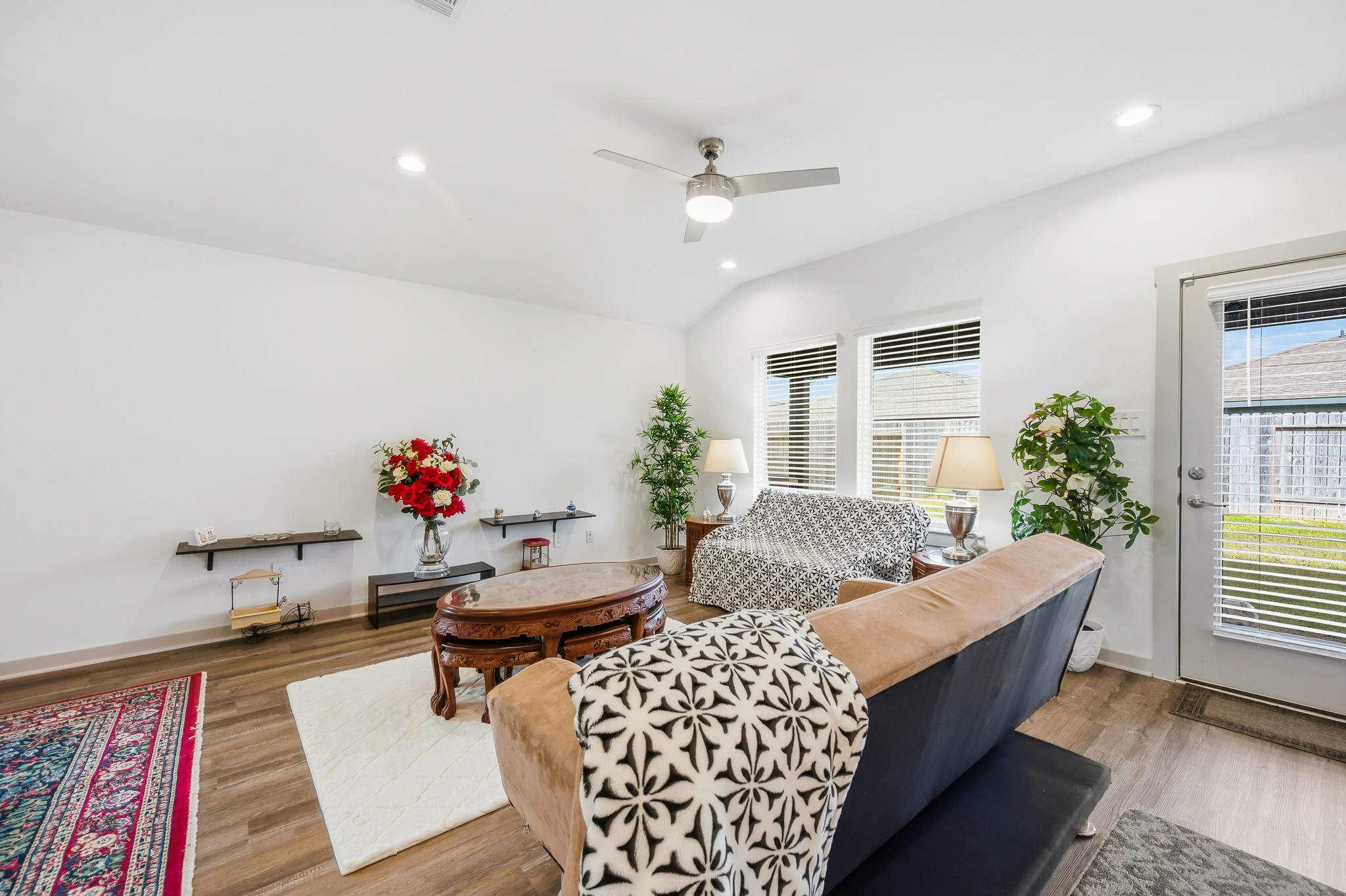 Bright and inviting living room with modern ceiling fan, large windows with blinds, and access to the backyard. Features wood flooring, cozy seating, and elegant decor with plants and a stylish rug.