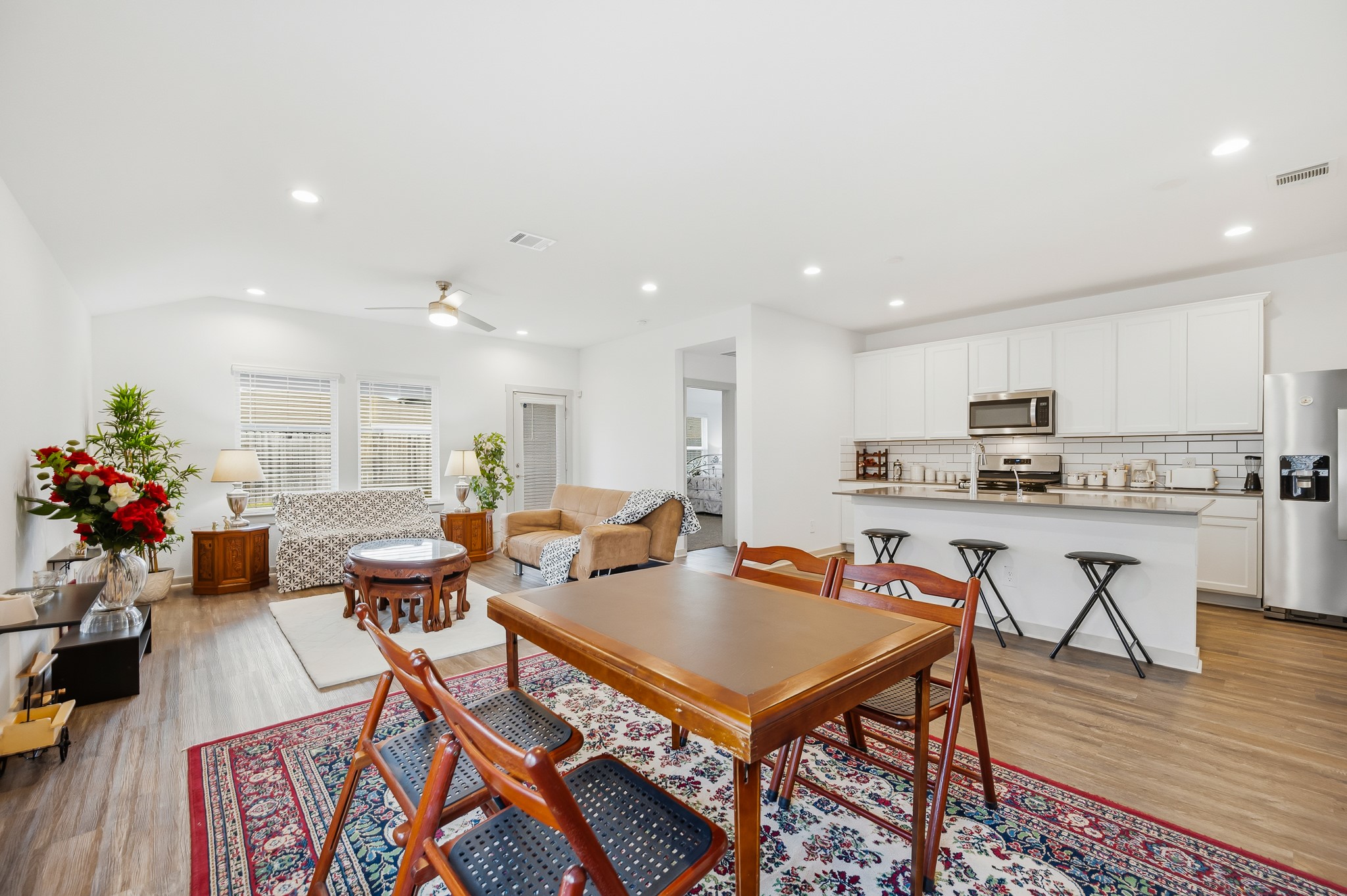Bright open-concept living area with hardwood floors, featuring a modern kitchen with white cabinets and stainless steel appliances. The space includes a cozy lounge area, dining table, and decorative elements like rugs and plants, creating a warm, inviting atmosphere.