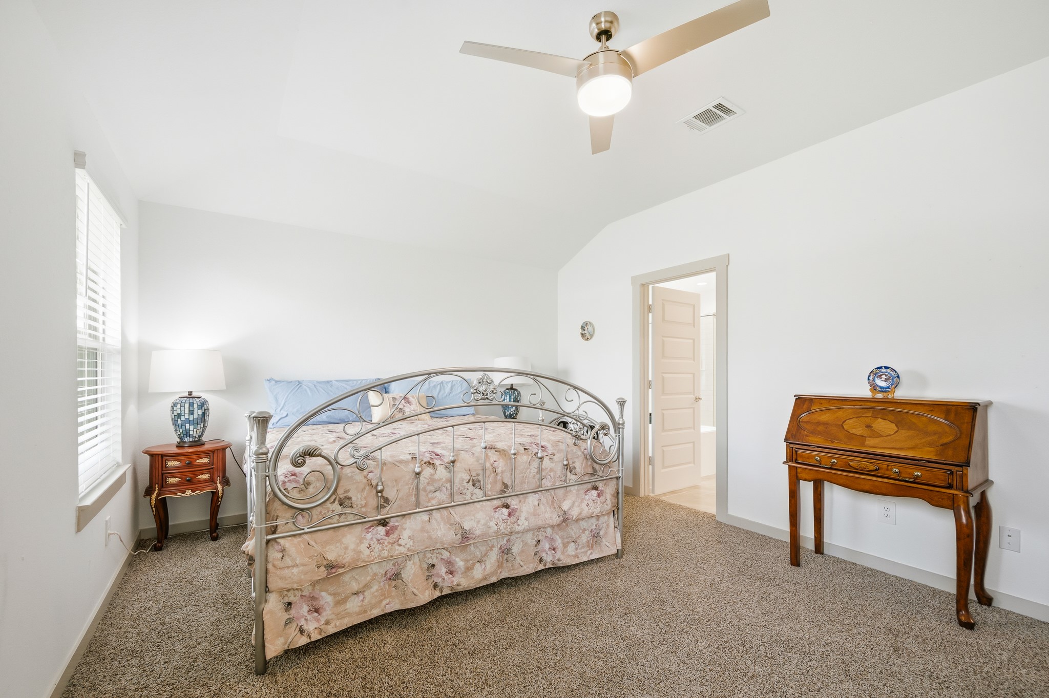 This bedroom features a cozy setting with neutral walls and carpet, a decorative metal bedframe, and ample natural light from a large window. A ceiling fan adds comfort, while a wooden side table and desk offer classic charm.
