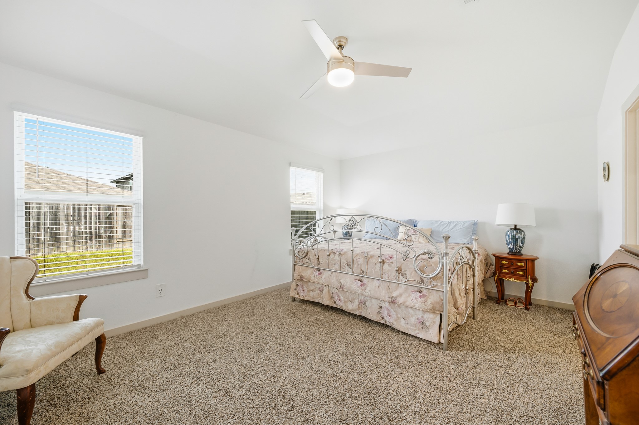 This bright bedroom features neutral walls and carpet, a ceiling fan, and ample natural light from two windows. It includes a decorative metal bed and classic furnishings, creating a cozy and inviting atmosphere.