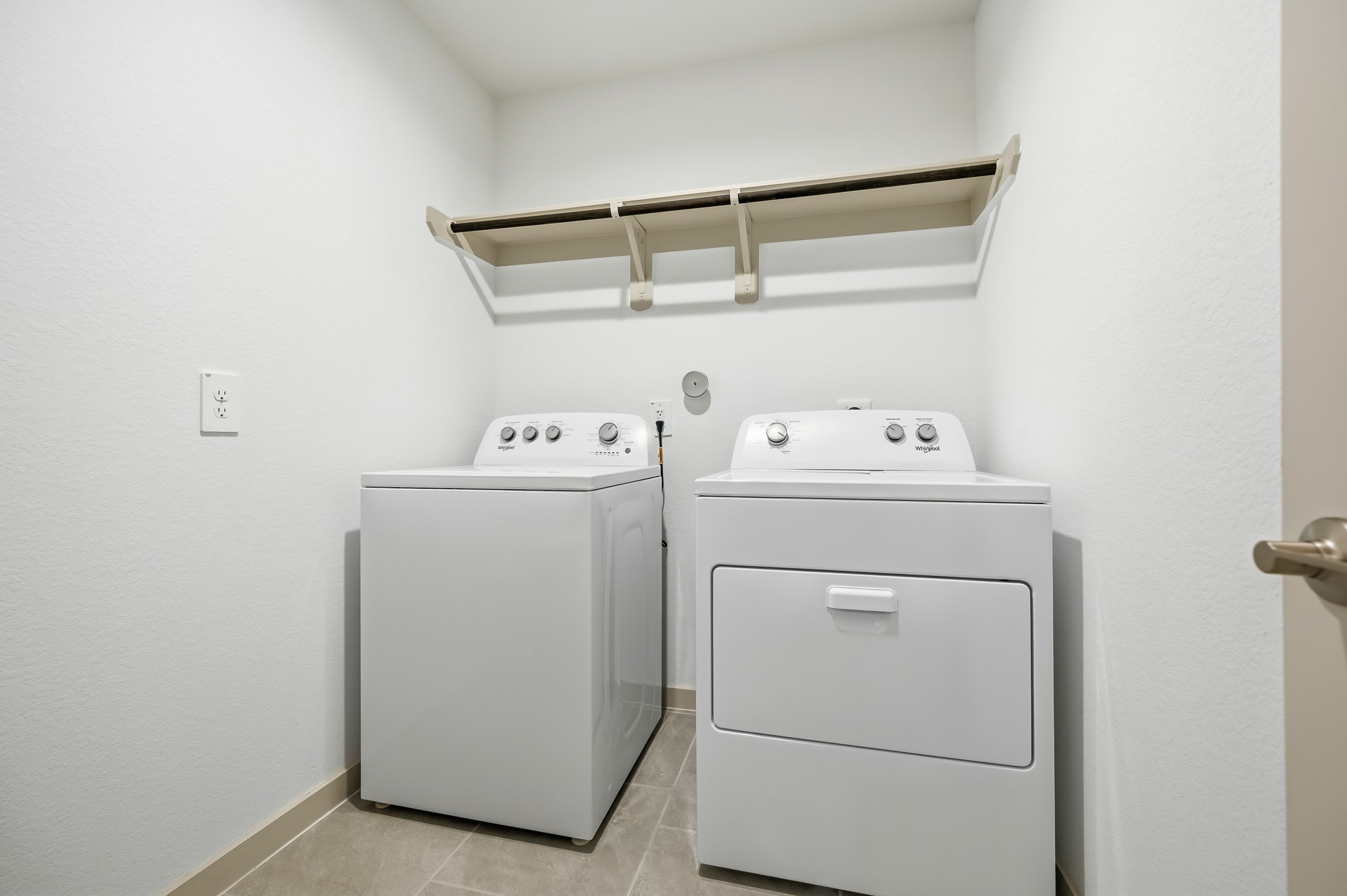 This photo shows a compact laundry room with a washer and dryer set, neutral walls, tile flooring, and a shelf for storage.