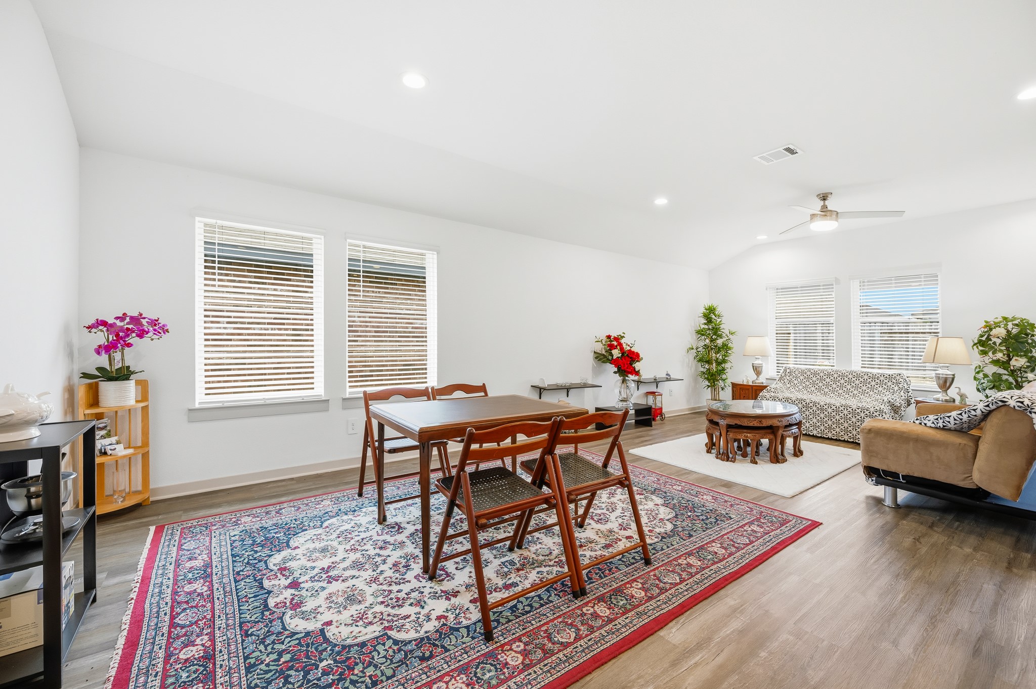 Bright and spacious living area with hardwood floors, featuring a dining space and cozy seating area. Large windows provide plenty of natural light, complemented by modern lighting and ceiling fan. Decorated with colorful rugs and plants for a welcoming atmosphere.