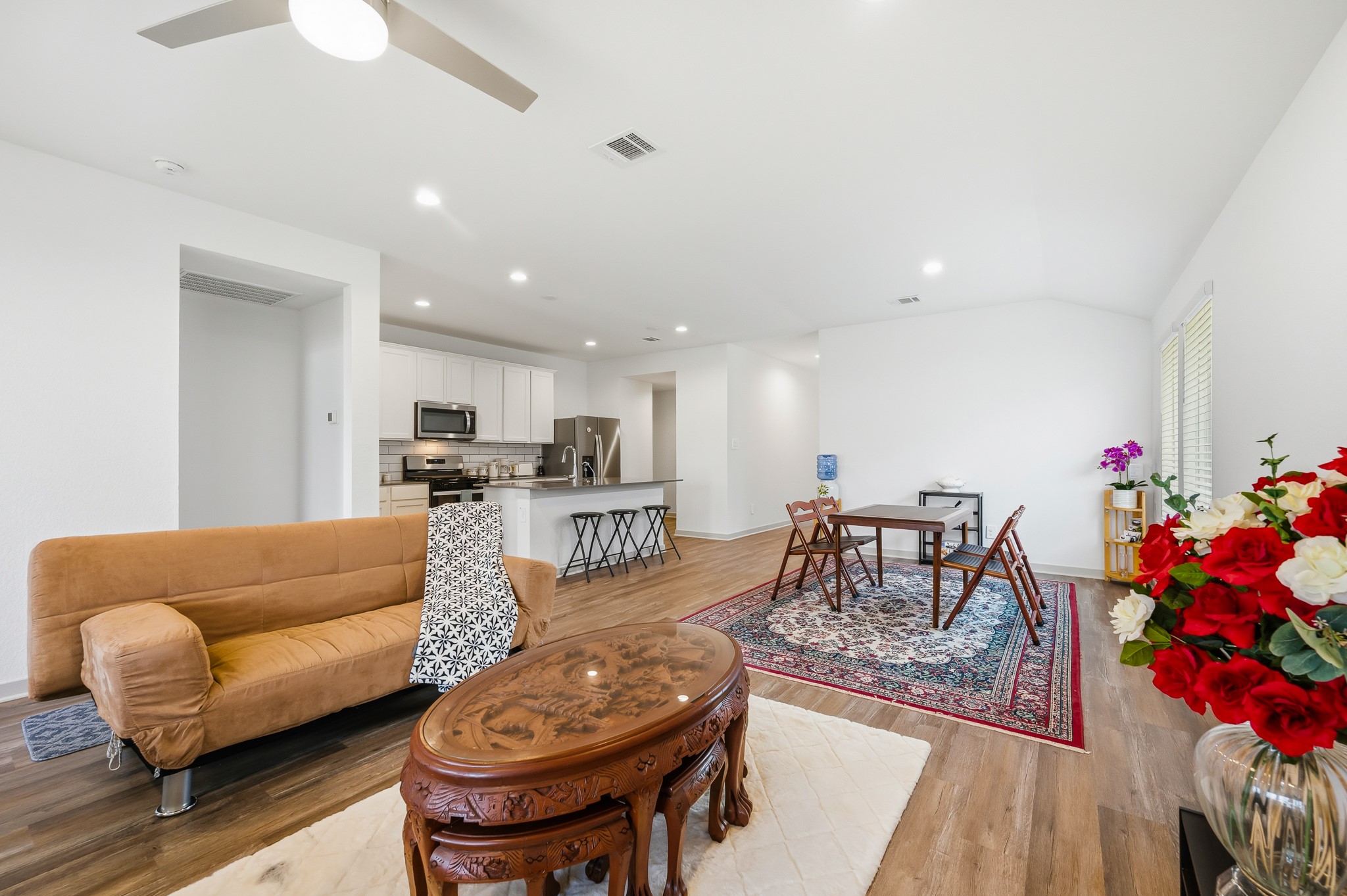 Bright and spacious open-concept living area with modern kitchen featuring stainless steel appliances. The room is accented by hardwood floors, a ceiling fan, and ample natural light. Cozy seating and a dining area with a decorative rug complete the space.
