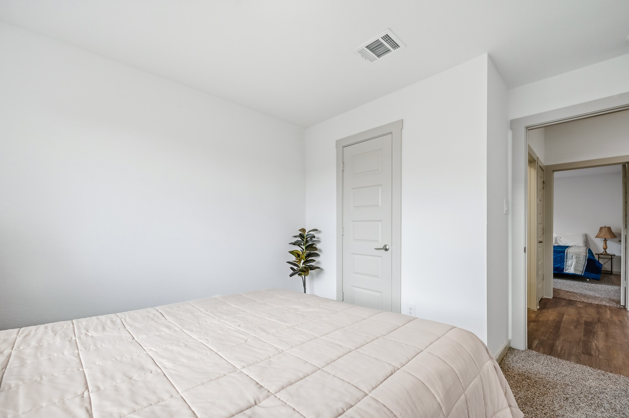This photo showcases a bright, minimalist bedroom with neutral walls and carpet. It features a neatly made bed, a small indoor plant, and a door leading to another room, creating an open feel.