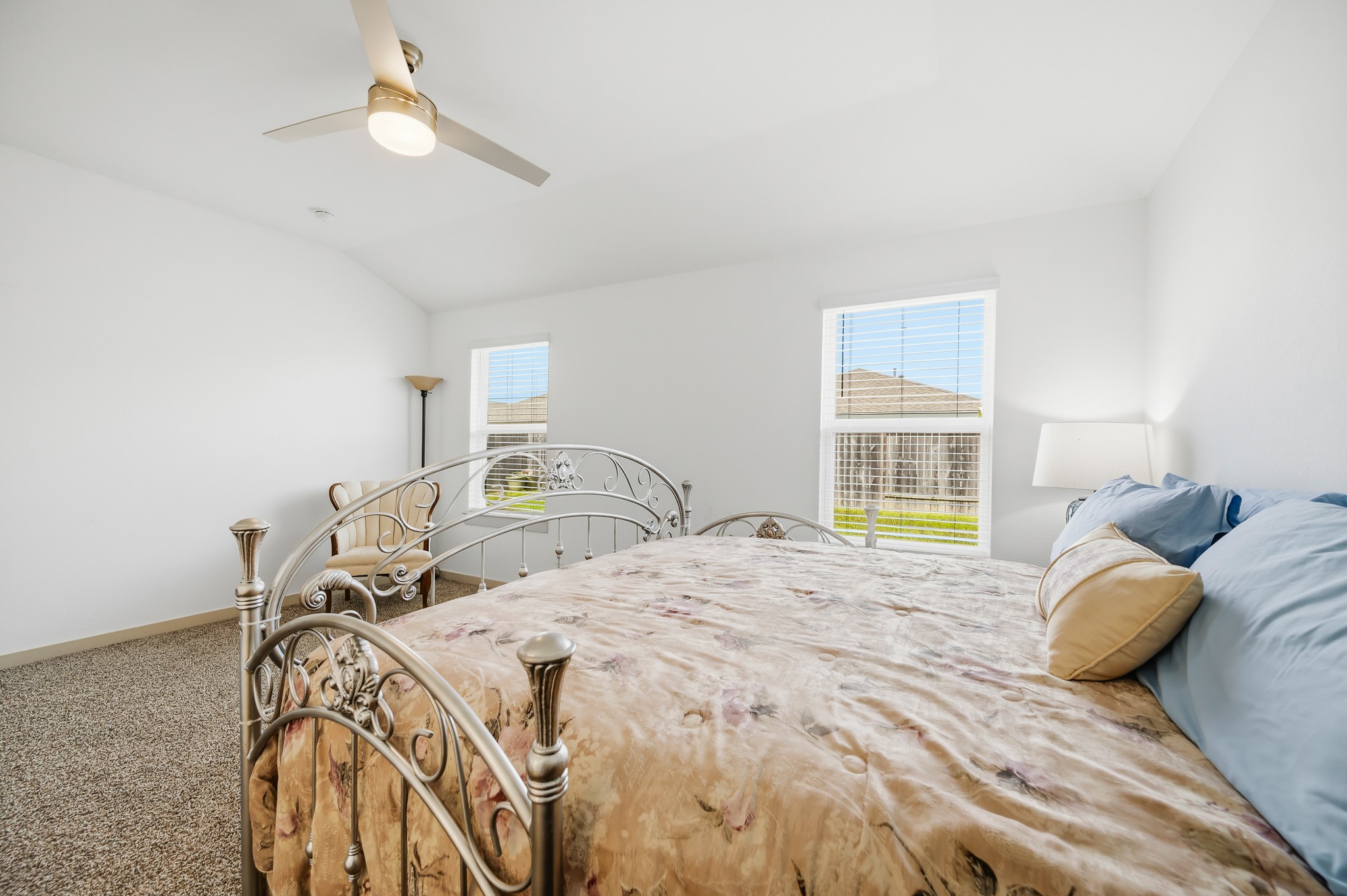 This bright bedroom features a cozy carpeted floor, stylish metal bed frame, and large windows that let in ample natural light. The ceiling fan and neutral decor create a comfortable, airy atmosphere.