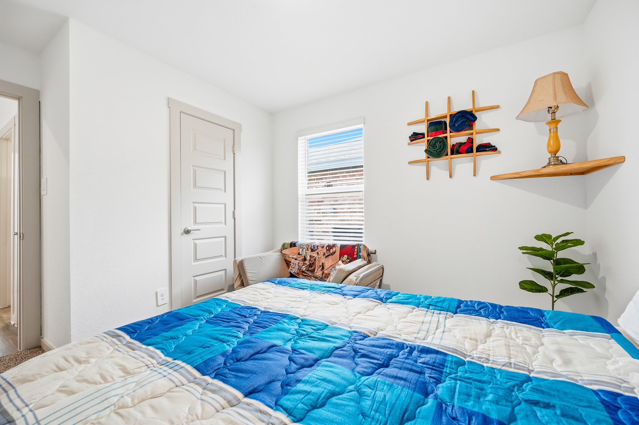 Cozy bedroom featuring a bright window, simple decor, and a colorful bedspread. The room includes a small shelf with a lamp and a decorative plant, creating a welcoming and functional space.