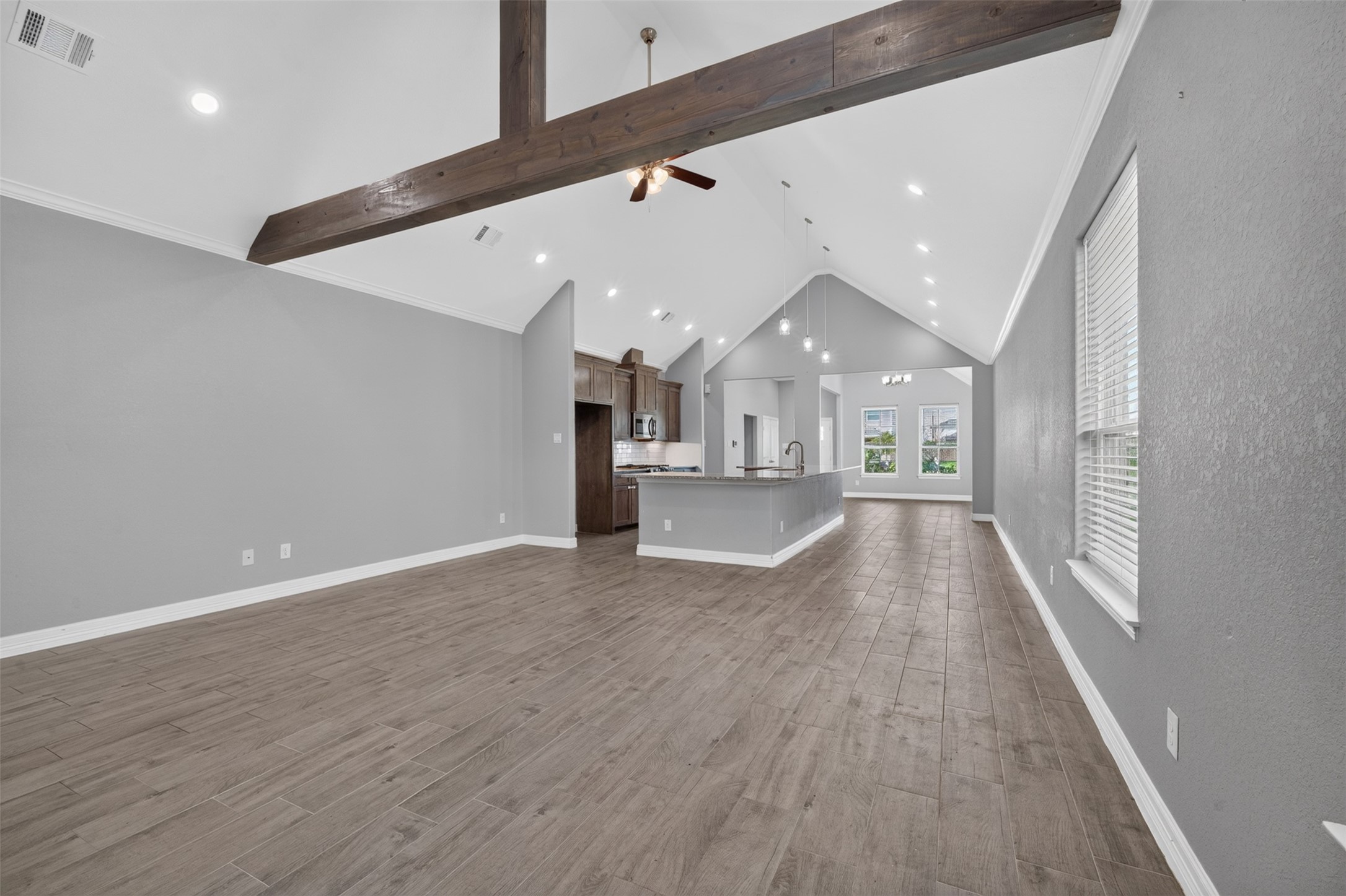 Spacious open-concept living area with a vaulted ceiling and exposed beam, featuring elegant wood-look tile flooring and abundant natural light from large windows.